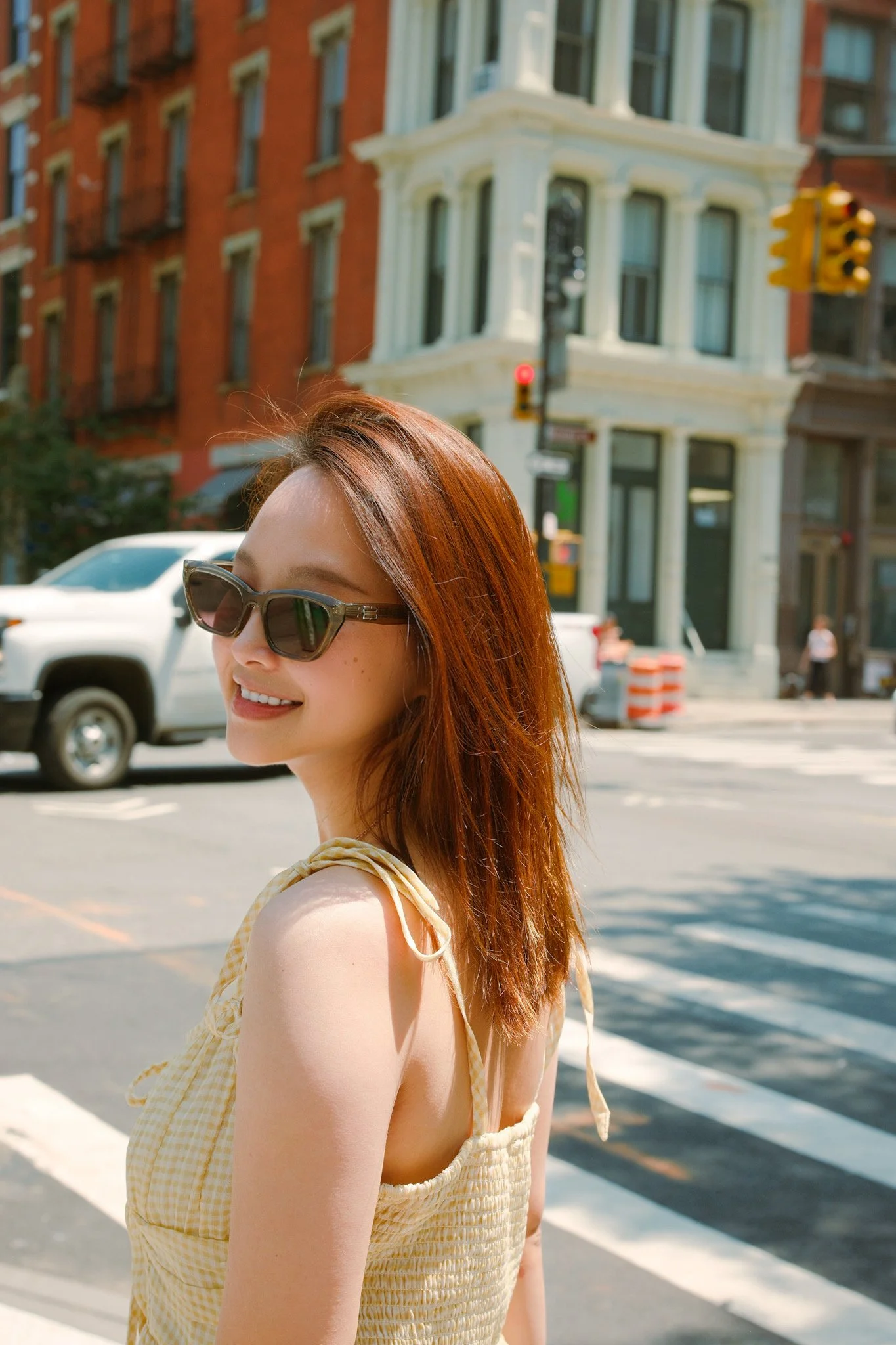 A young woman with reddish-brown hair and sunglasses smiling on a city street. In the background, there are buildings, cars, and a traffic light.