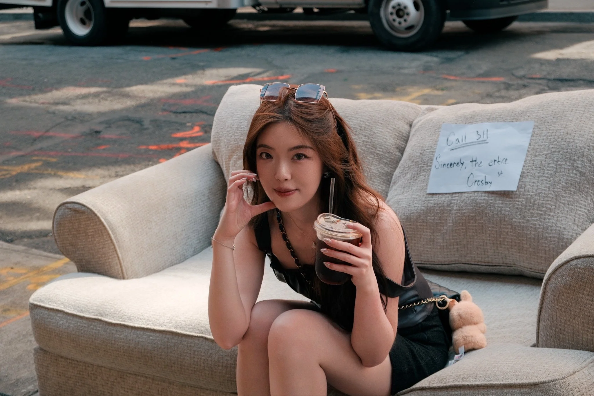 A woman with long brown hair, sunglasses on her head, and wearing a black sleeveless top is sitting on a beige couch outdoors. She is holding a cup of iced coffee and smiling at the camera. There is a piece of paper with writing pinned to the couch b