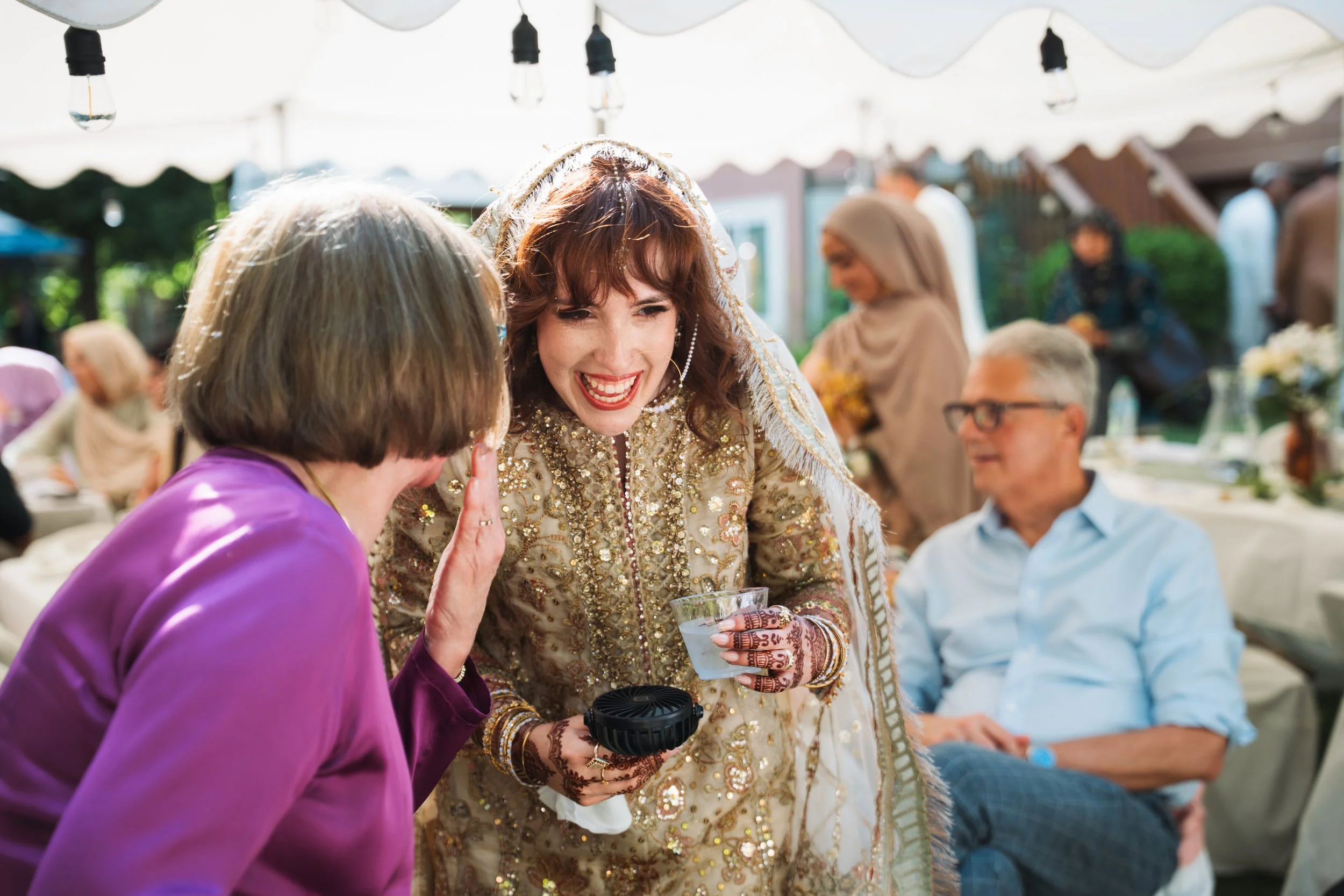 A woman in traditional South Asian attire smiling and talking to another woman in a purple outfit at an outdoor gathering with string lights overhead.