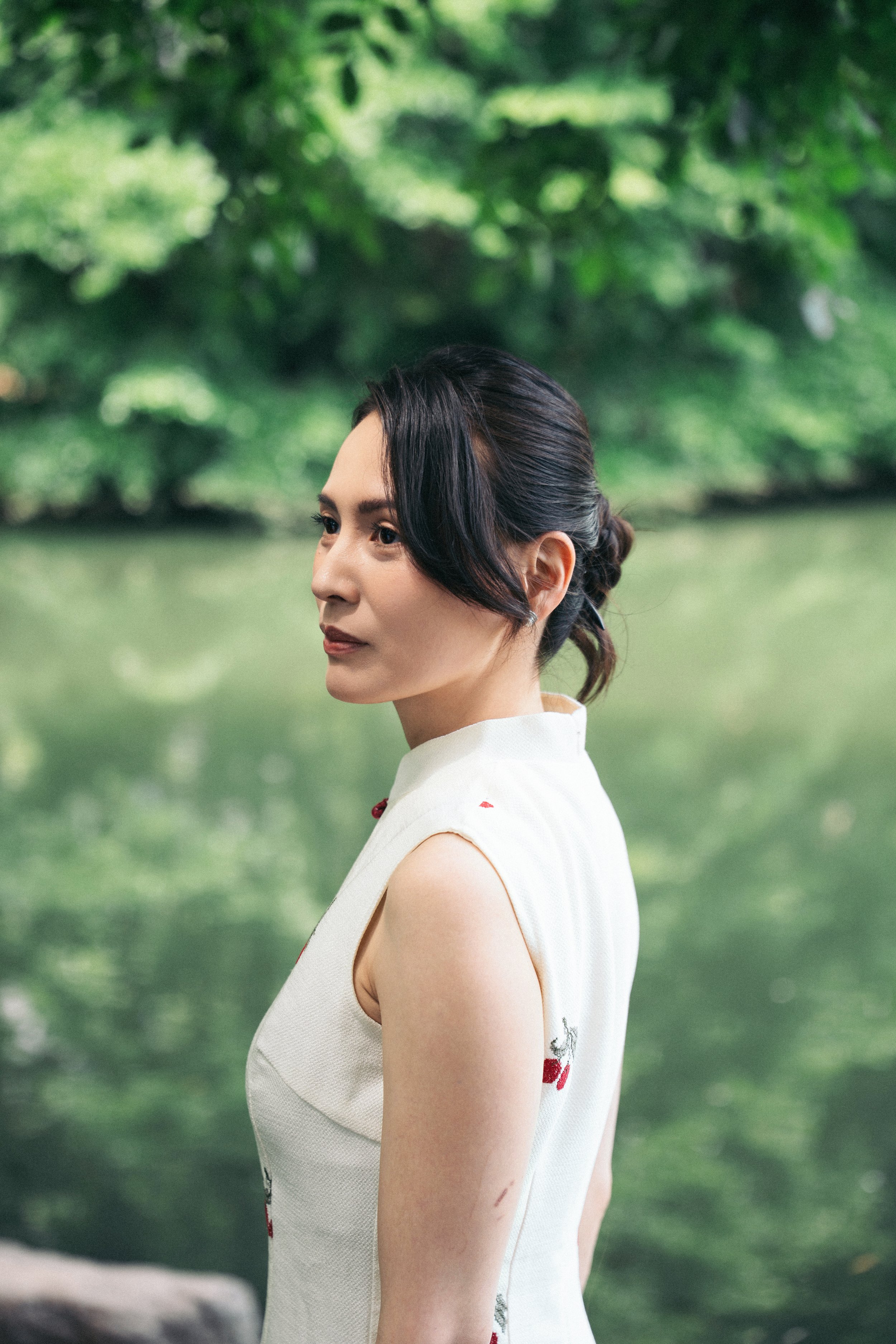 A woman with dark hair styled in an elegant updo, wearing a sleeveless white dress with floral embroidery, stands outdoors near a body of water with green foliage in the background.