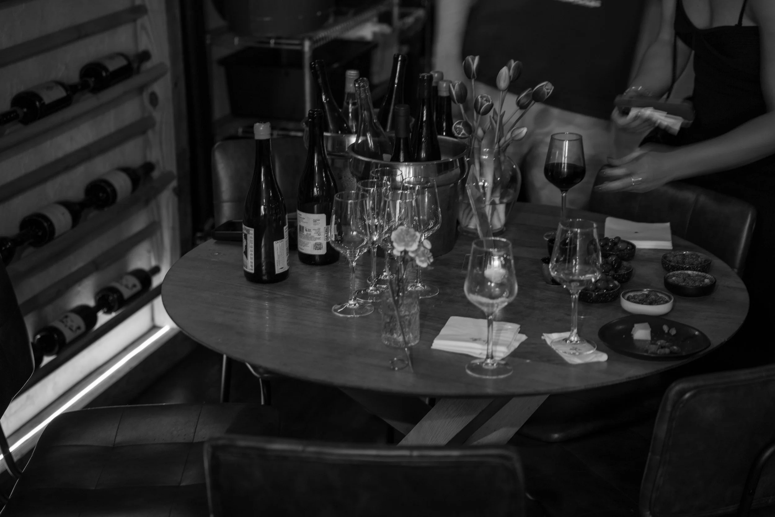 A table set for a gathering with wine bottles, glasses, tulips, and small bowls of snacks or dips, with a wine rack in the background.