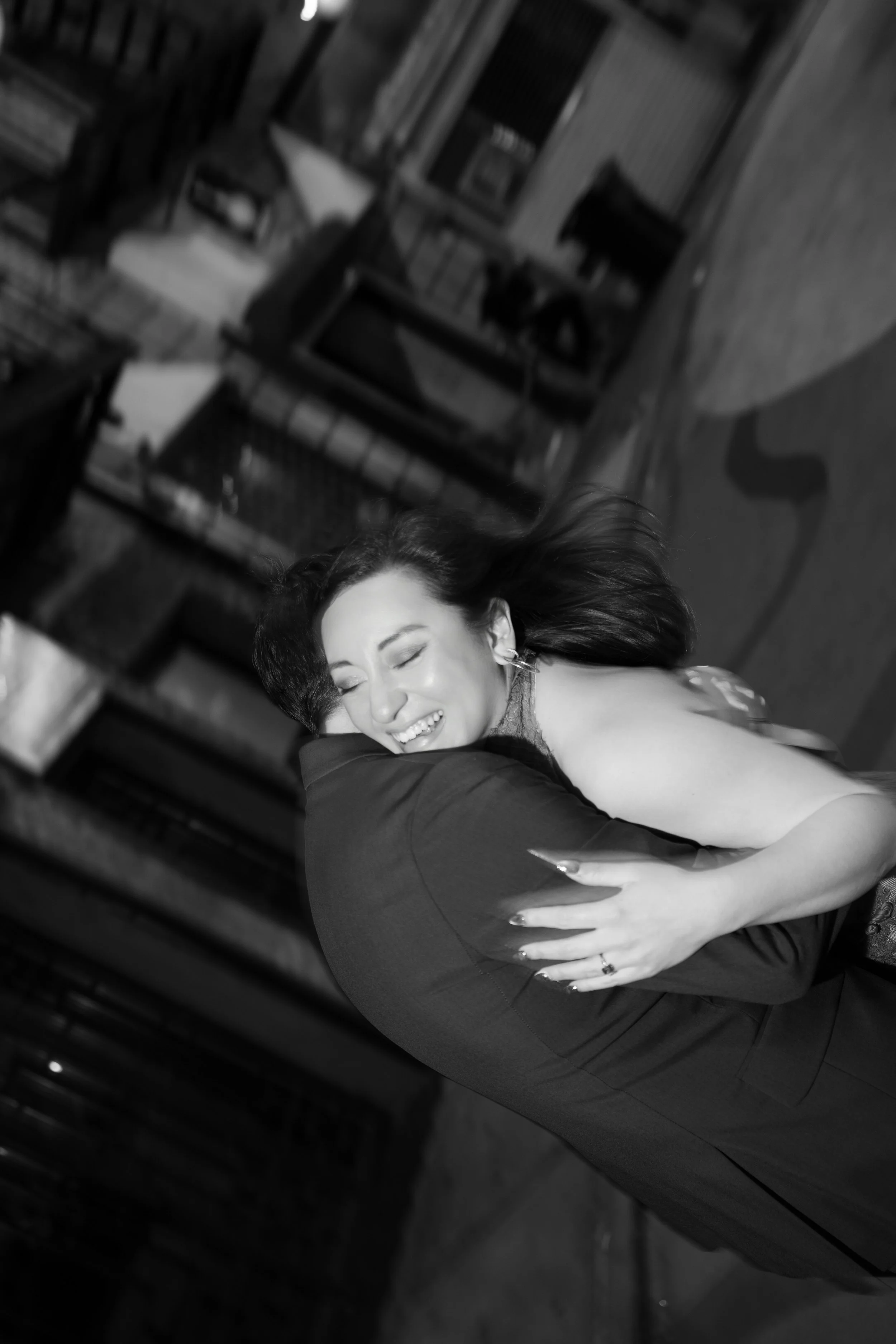 A woman with dark hair smiling and embracing someone, possibly at a dance or social event, in a room with wooden furniture and a man in the background.