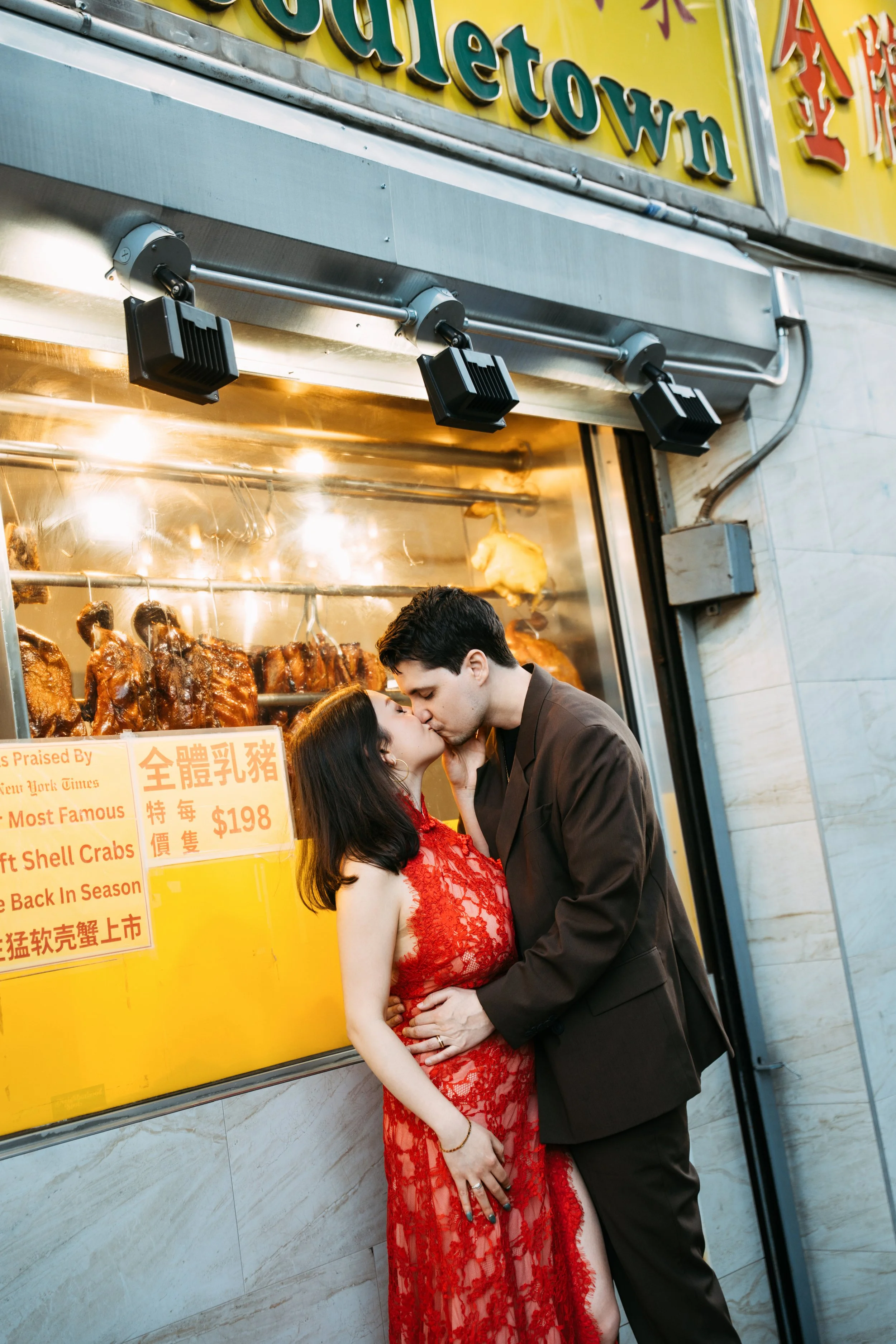 A couple sharing a kiss in front of a neon-lit food stand with hanging roasted meats and a yellow sign with Chinese and English text.