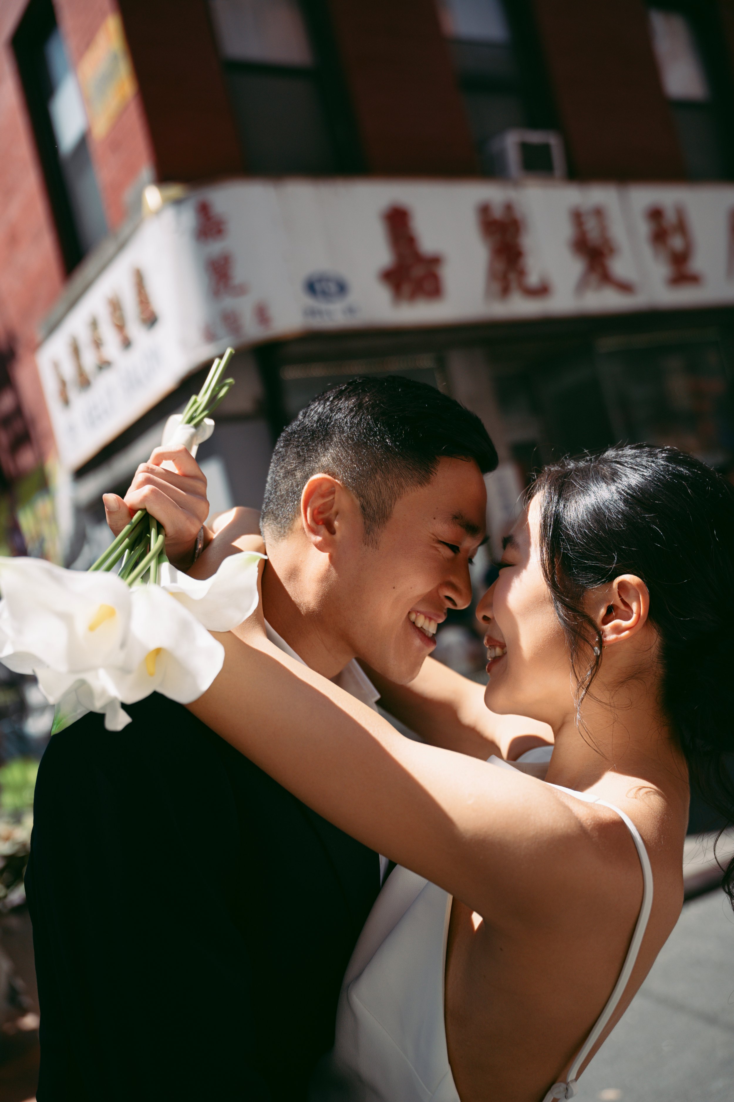 A close-up of a newlywed couple smiling and touching foreheads during their wedding celebration outdoors, with the groom holding white flowers.