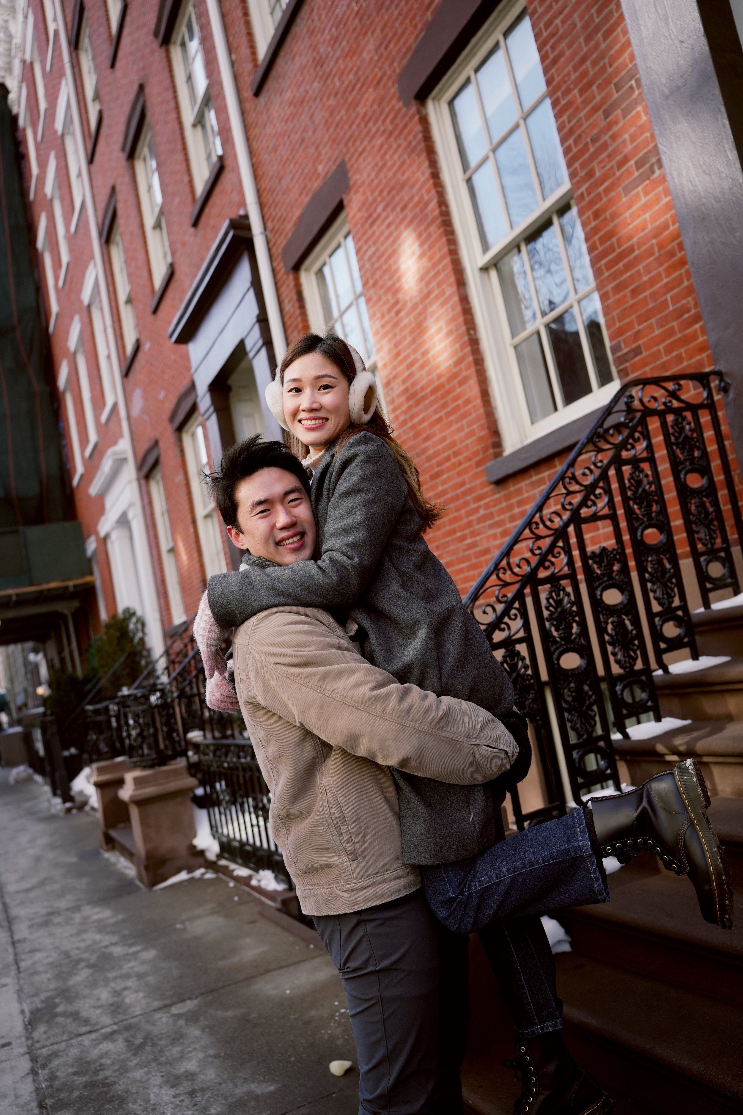 A smiling young man lifting a young woman in a warm coat and earmuffs in front of a red brick residential building with large windows and black wrought iron stairs, winter scene.