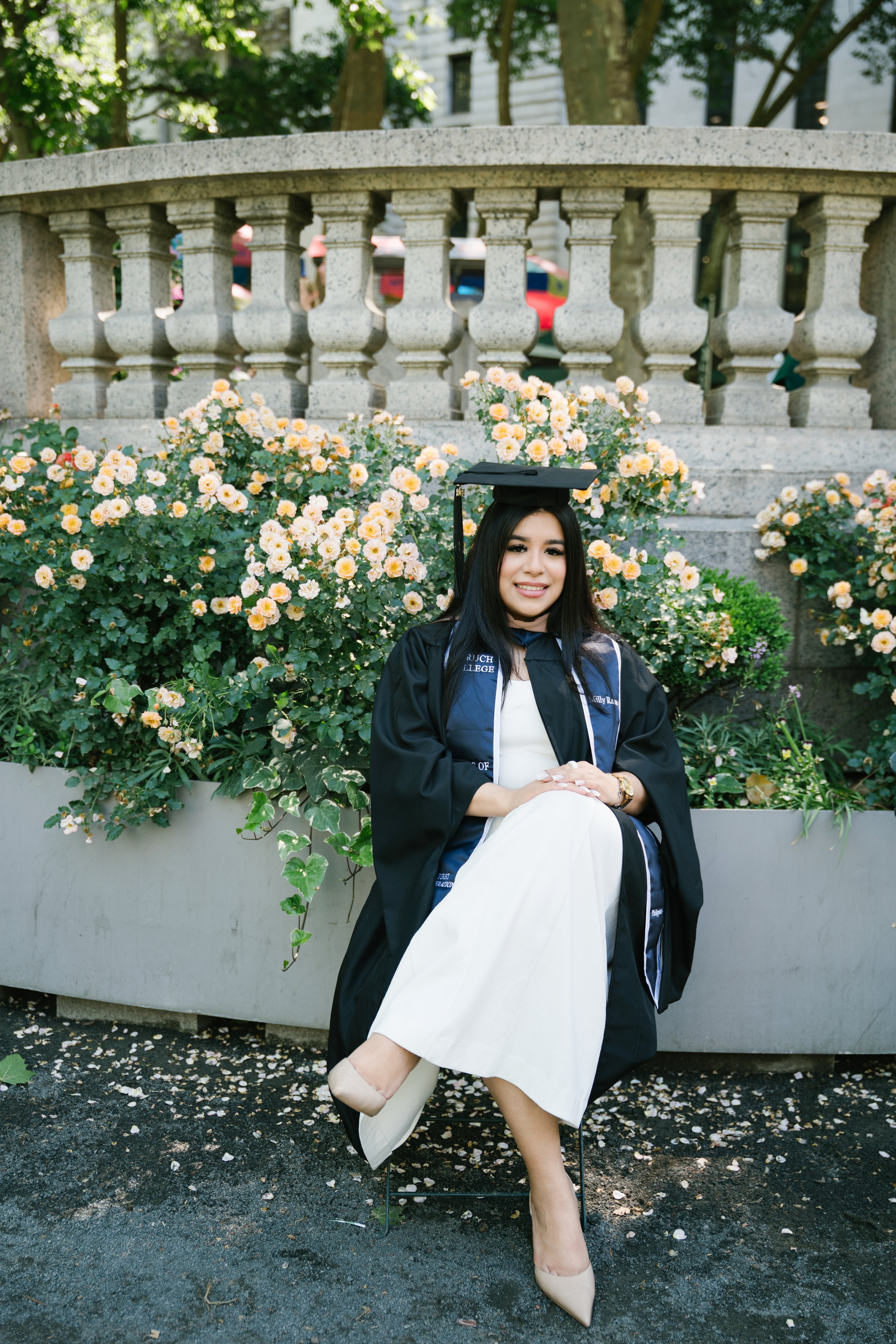 A young woman in a black graduation gown and cap, sitting outdoors in front of a flowerbed of yellow roses, smiling at the camera.