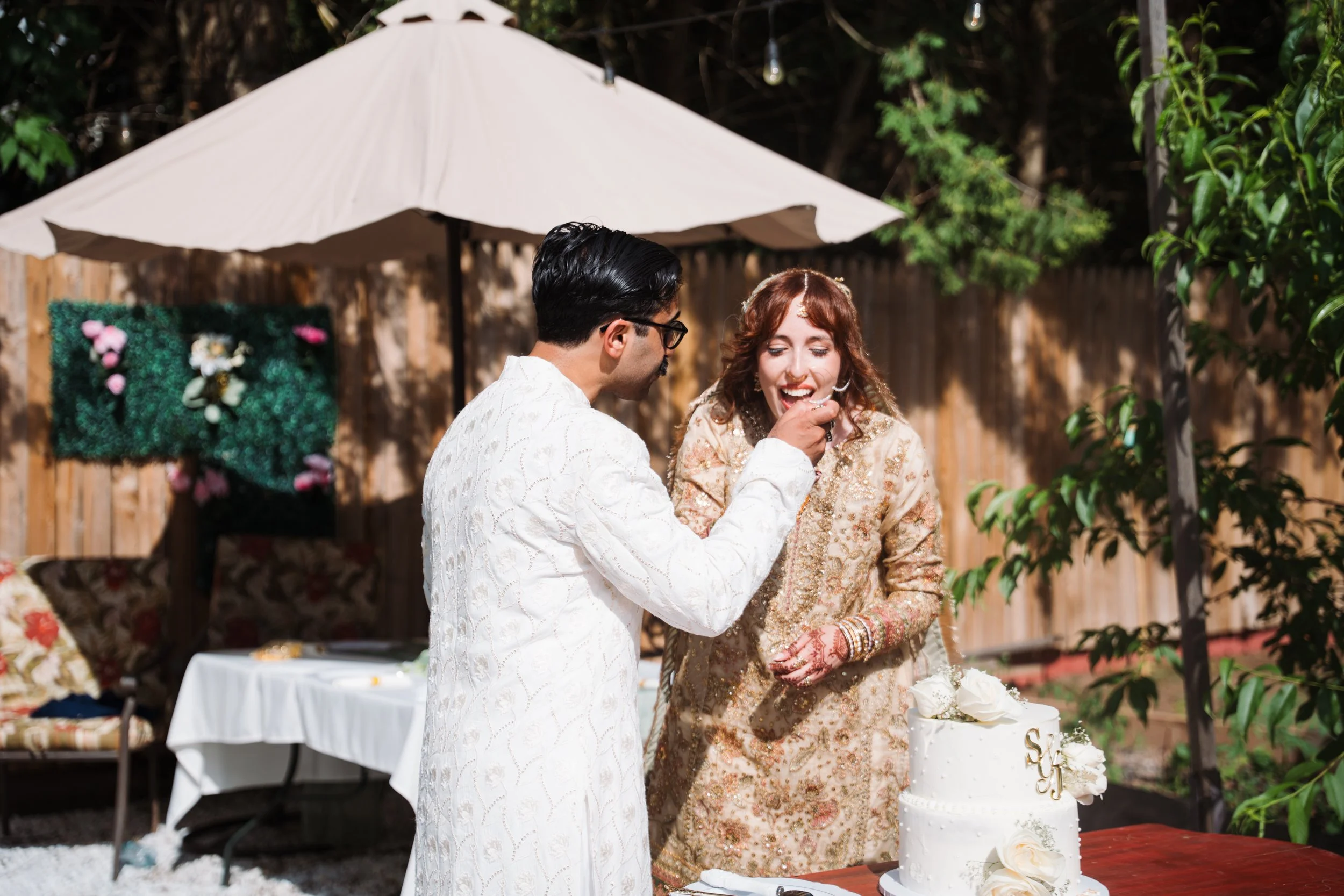 A couple at an outdoor wedding, with the man in a white traditional outfit and the woman in a gold embroidered dress, cutting a white wedding cake decorated with gold initials.