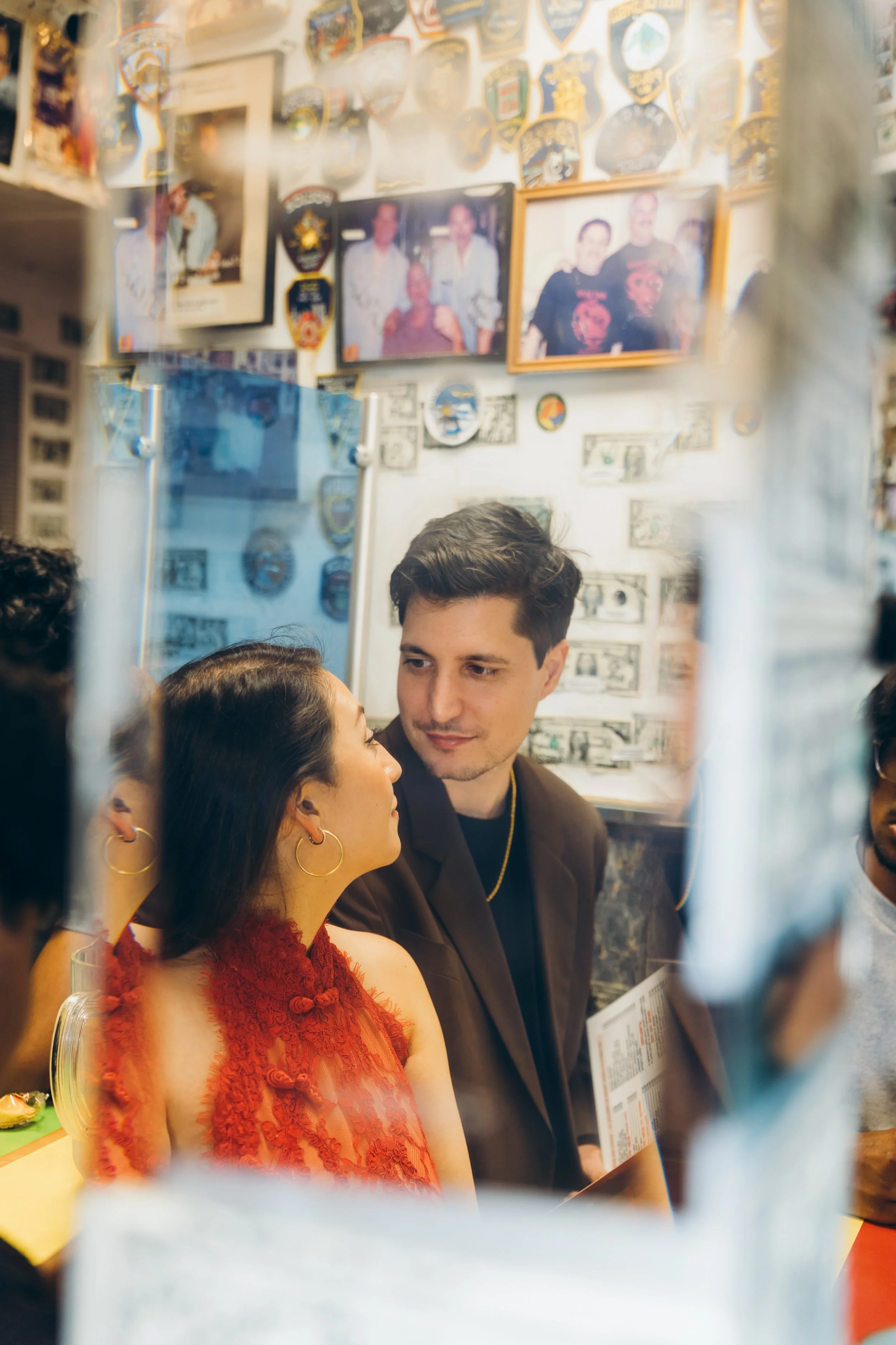 A couple staring at each other inside a restaurant, with the woman's face turned towards the man's face. The scene is viewed through a reflective surface with various framed pictures and patches on the wall behind them, including some money on the wa