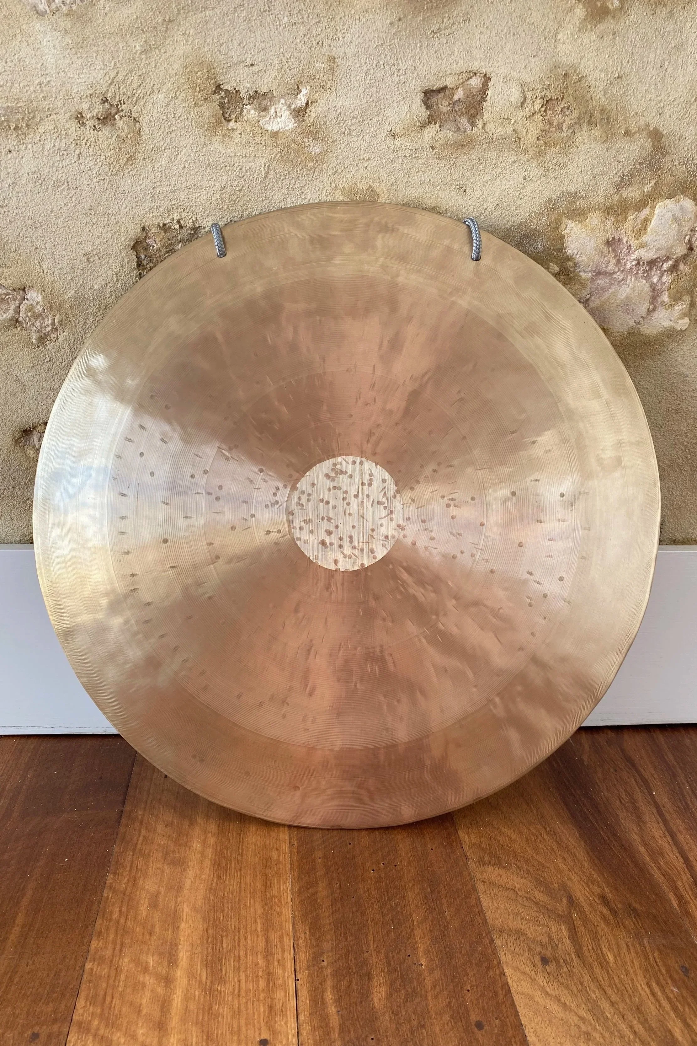 Brass-coloured therapeutic gong on wooden floor, leant against stone wall