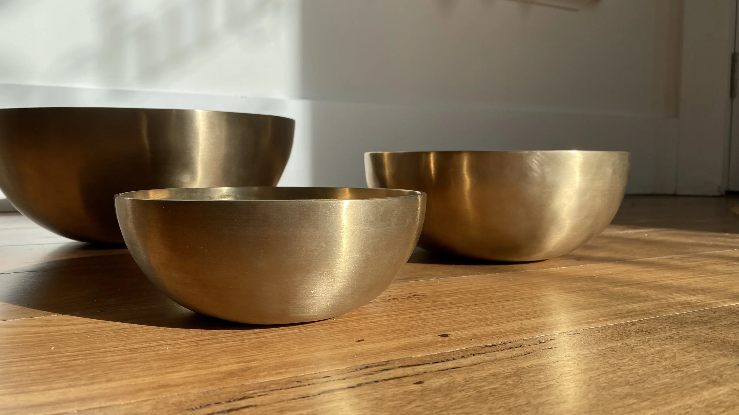 Brass-coloured therapeutic sound bowls of various sizes on wooden floor in the sun