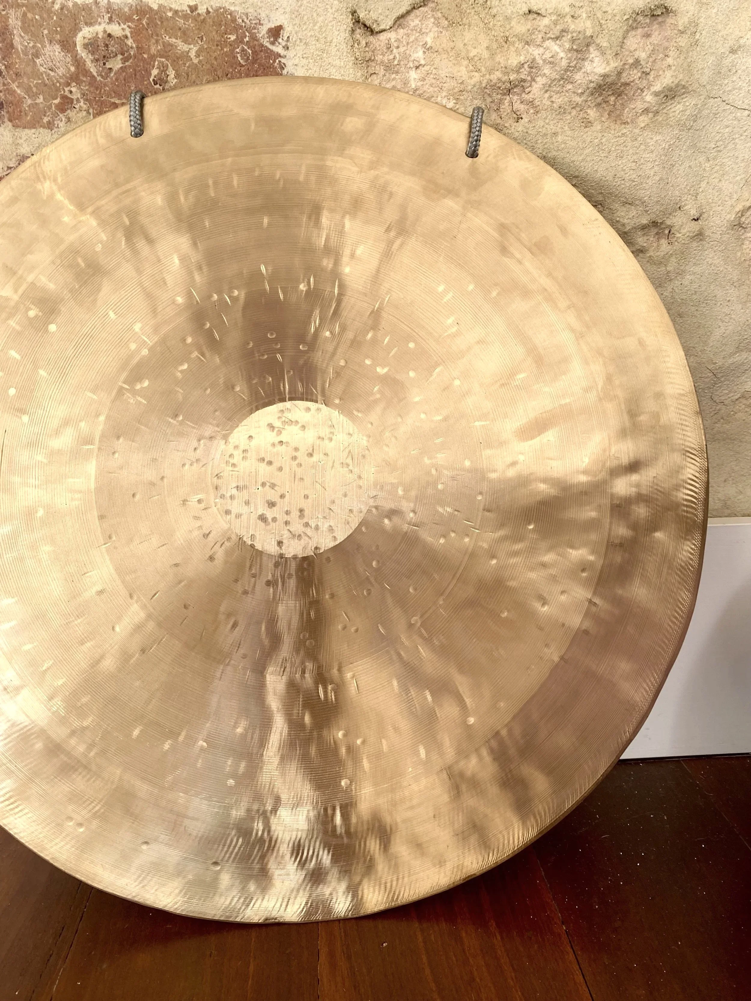 Brass-coloured therapeutic gong on wooden floor, leant against stone wall