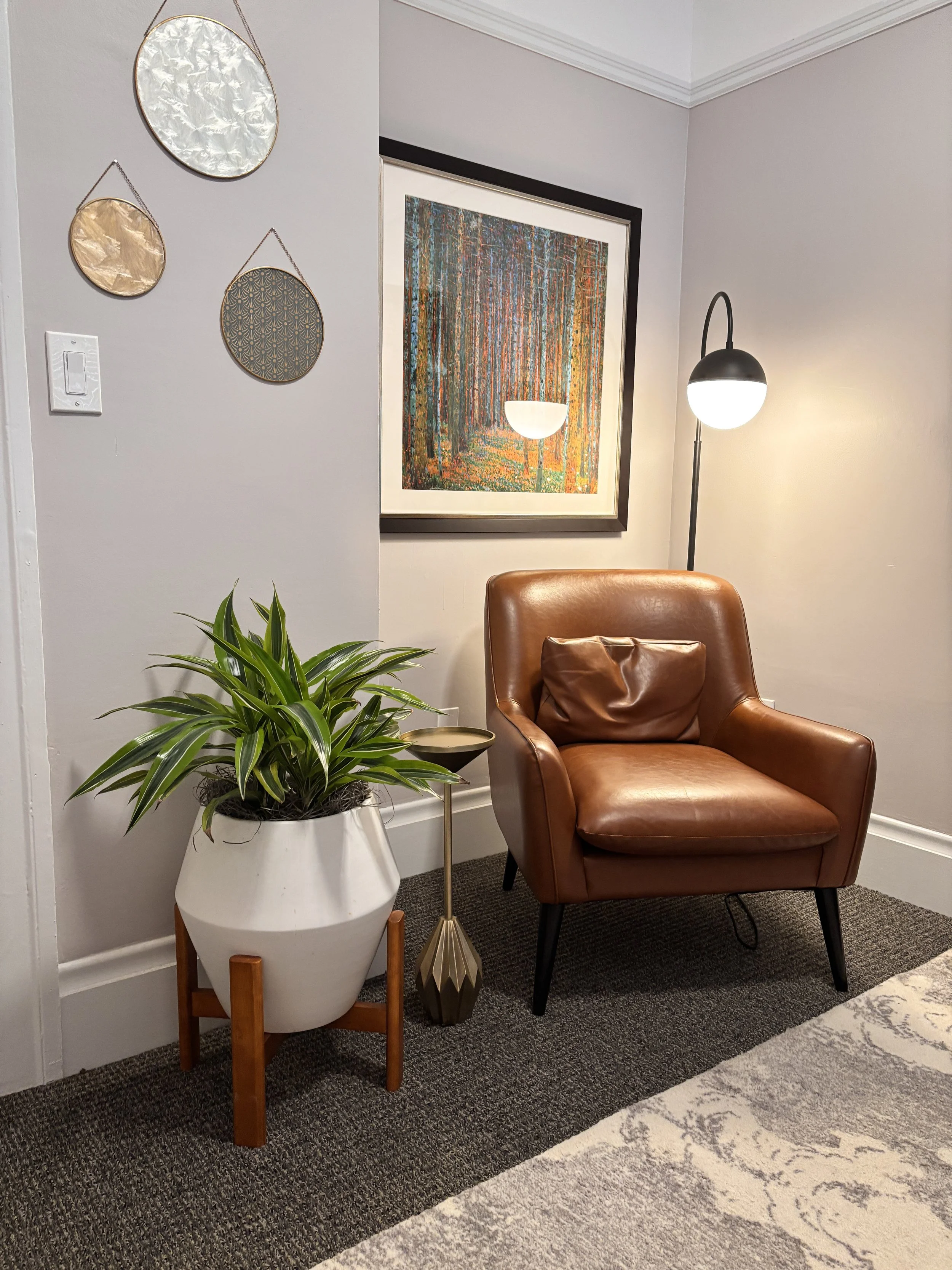 A cozy corner with a brown leather armchair, a tall black floor lamp with a round white shade, a white planter with a green houseplant on a wooden stand, pinkish-beige walls, a textured multicolored painting, and decorative wall hangings with circular, metallic designs.