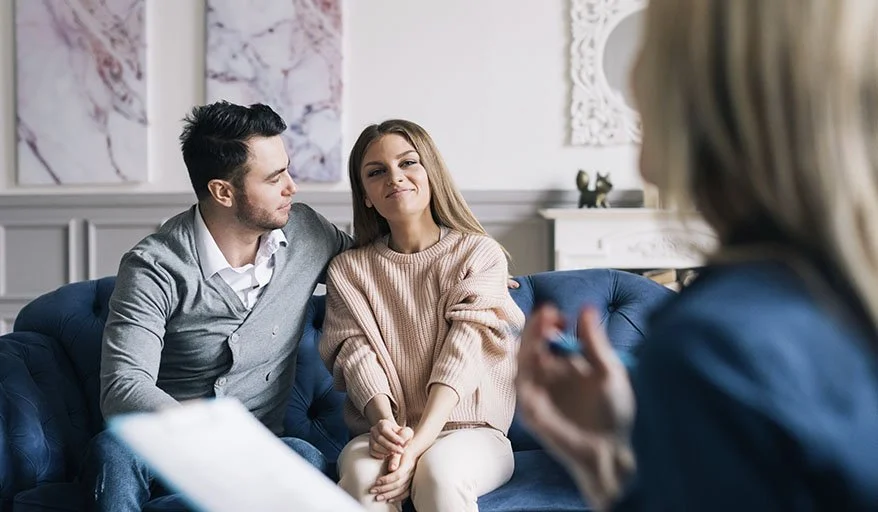 A couple sitting on a blue couch during a counseling session with a therapist, engaging in conversation.