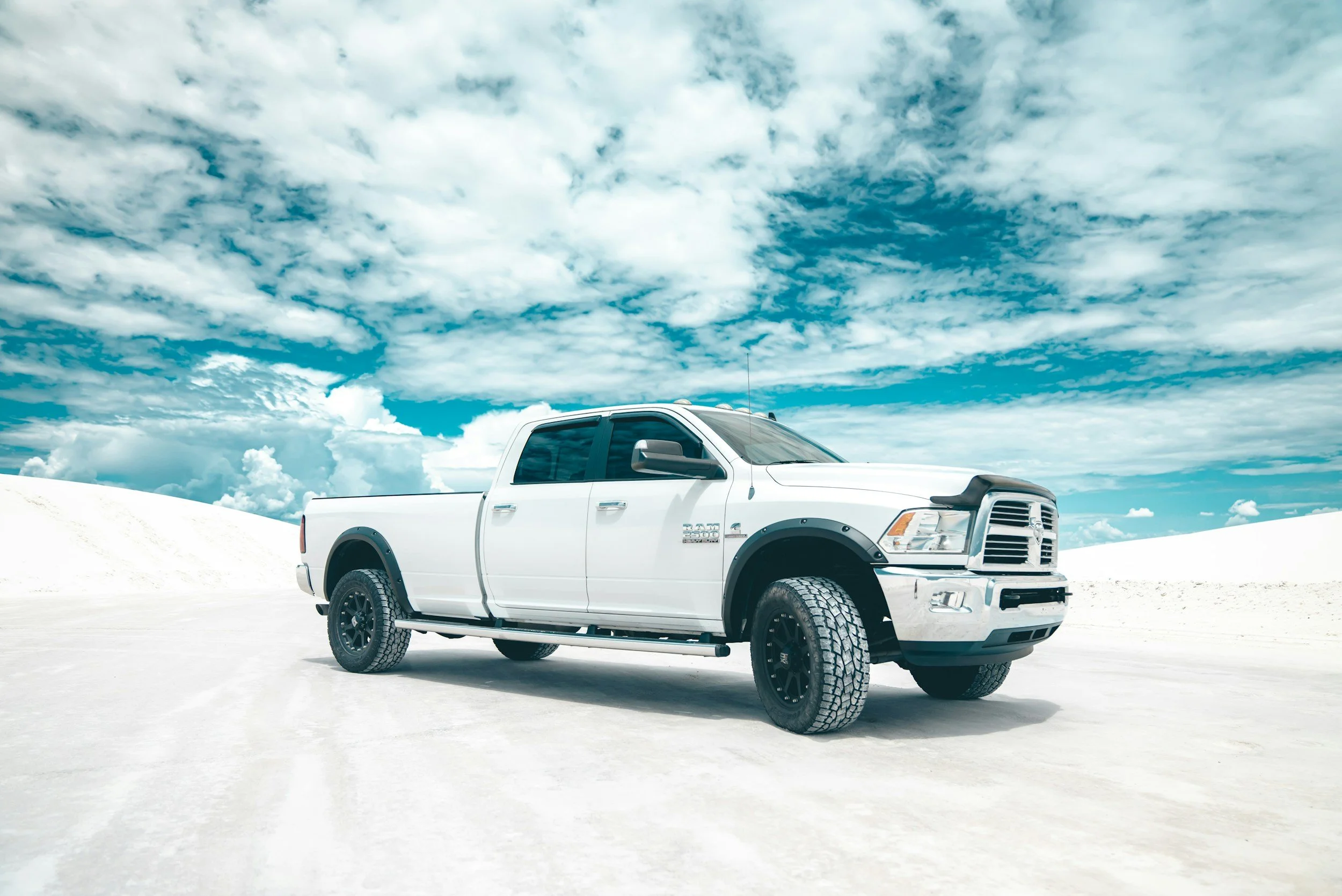 A white truck with black wheels parked on white sand against a backdrop of a partly cloudy blue sky.