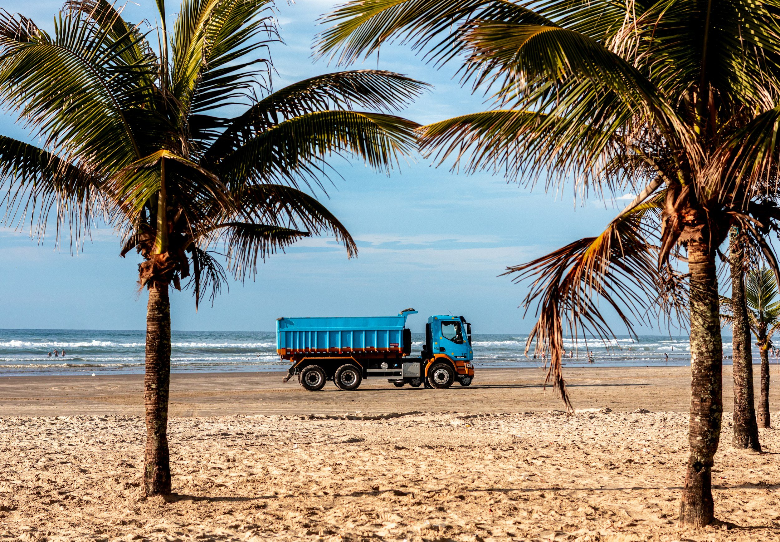 A beach scene with tall palm trees in the foreground, a blue truck on the sandy shore, and people in the water in the background. The sky is partly cloudy and the ocean waves are rolling ashore.