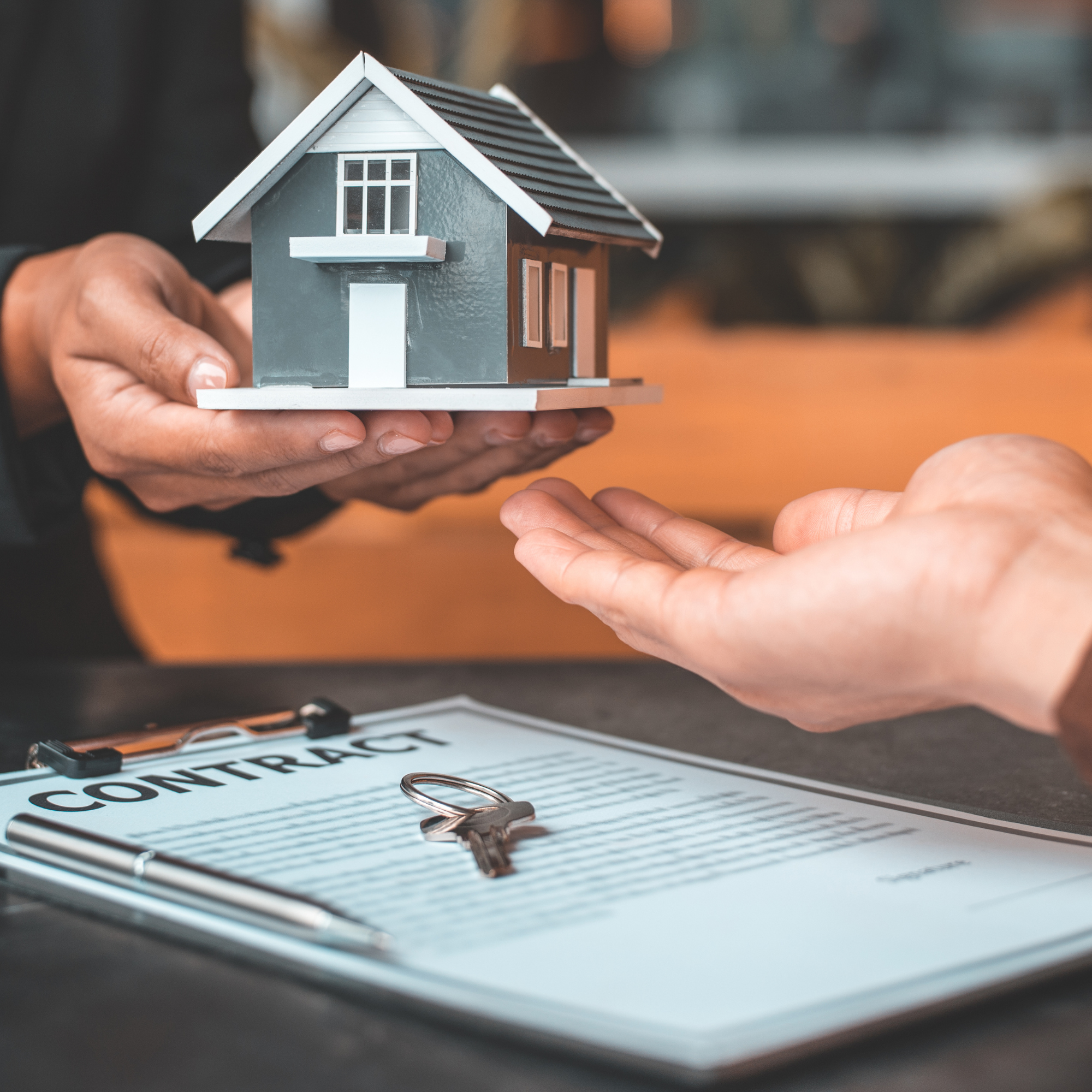 A person holding a small model house with dark gray walls, white window frames, and a brown roof. The model house is being handed over to another person's hand, which is positioned underneath it. In the background, there is a document with the word 'CONTRACT' visible, along with metal paperclips placed on top of it.
