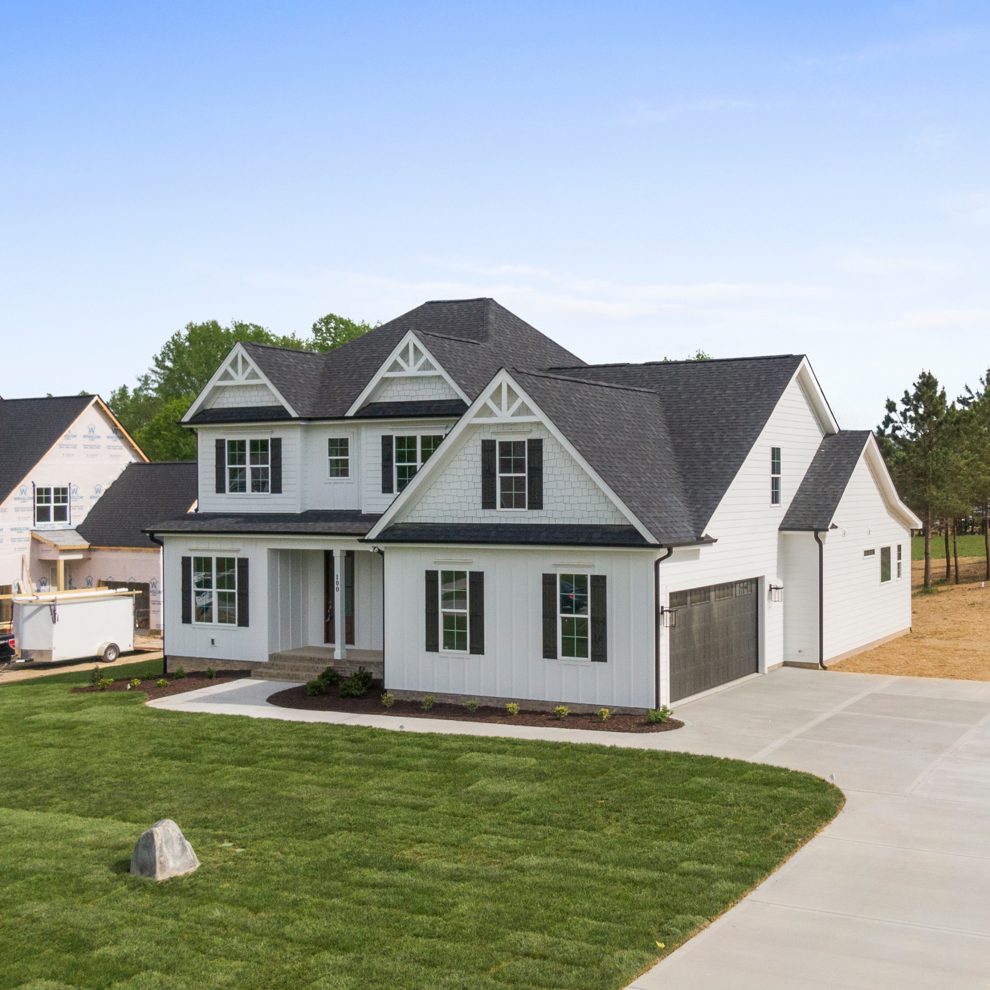 A white two-story house with black shutters and a dark shingle roof, surrounded by a green lawn and trees, under a blue sky.