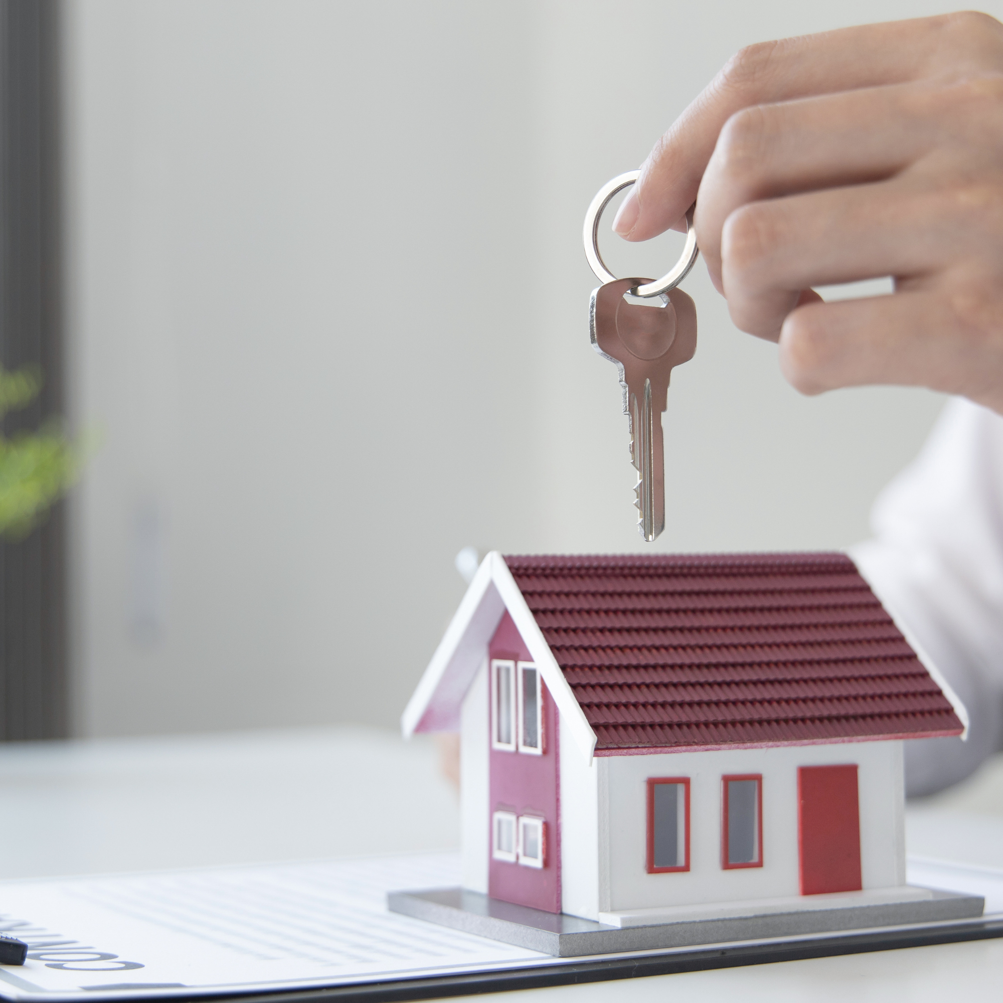 A miniature model of a pink house with a red roof and white trim, placed on a white surface, with a key hanging from a person's fingers above it.