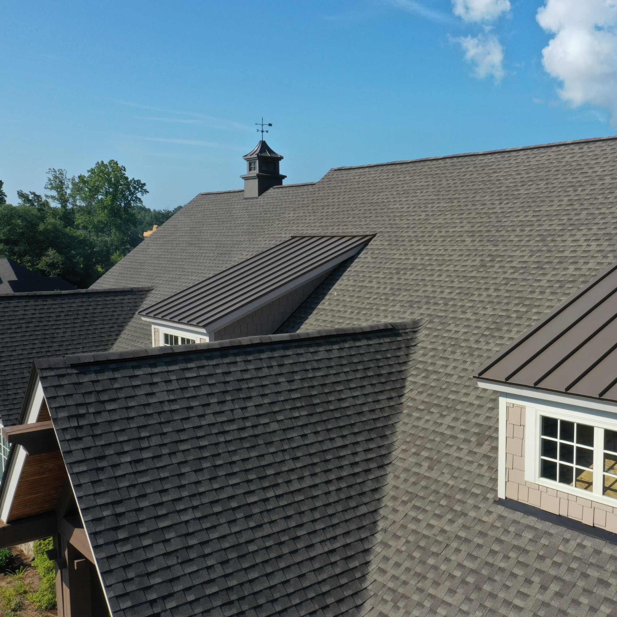 A man with a shaved head wearing a green shirt and jeans kneeling on a shingled roof, working with a clipboard, beneath a clear blue sky with some clouds and trees.