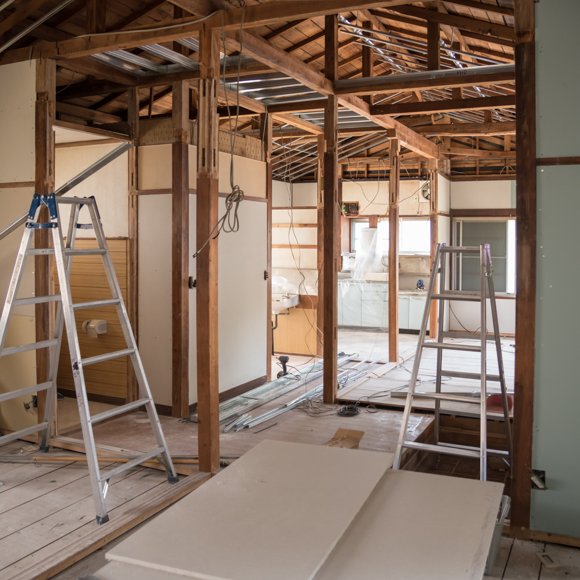 Interior view of a house under renovation with exposed wooden framing, metal roofing, ladders, electrical wiring, and construction materials.