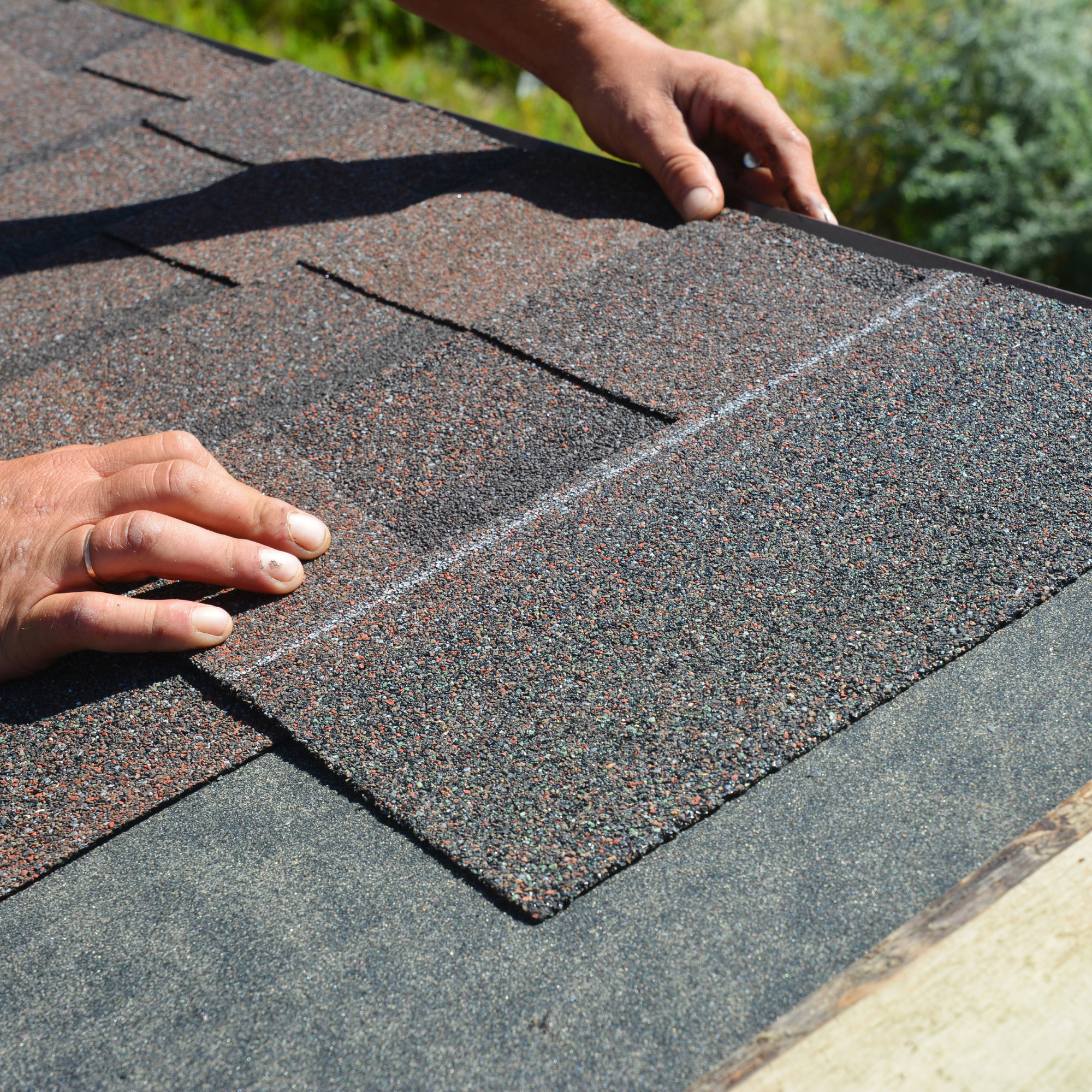 A person in a grey shirt and white gloves working on a roof with dark brown shingles, wearing a yellow safety helmet and a neon safety vest, using a hammer placed on the shingles.