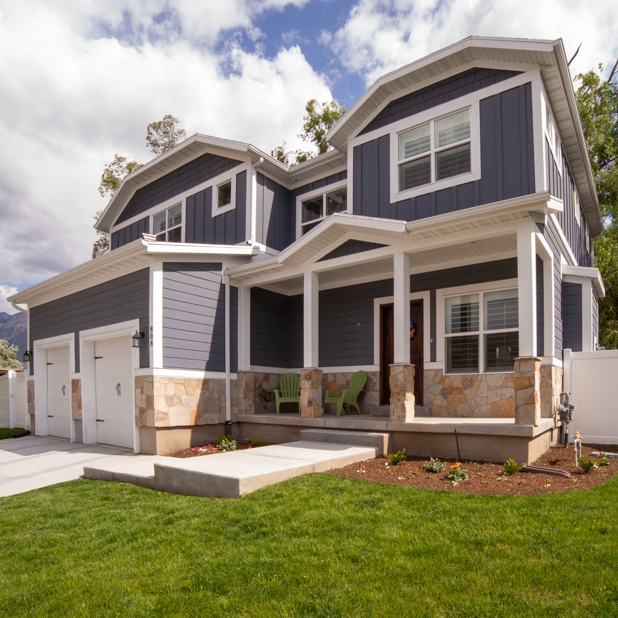 A modern two-story house with dark gray siding, large windows with white frames, a stone foundation, green lawn, and a porch with green Adirondack chairs.