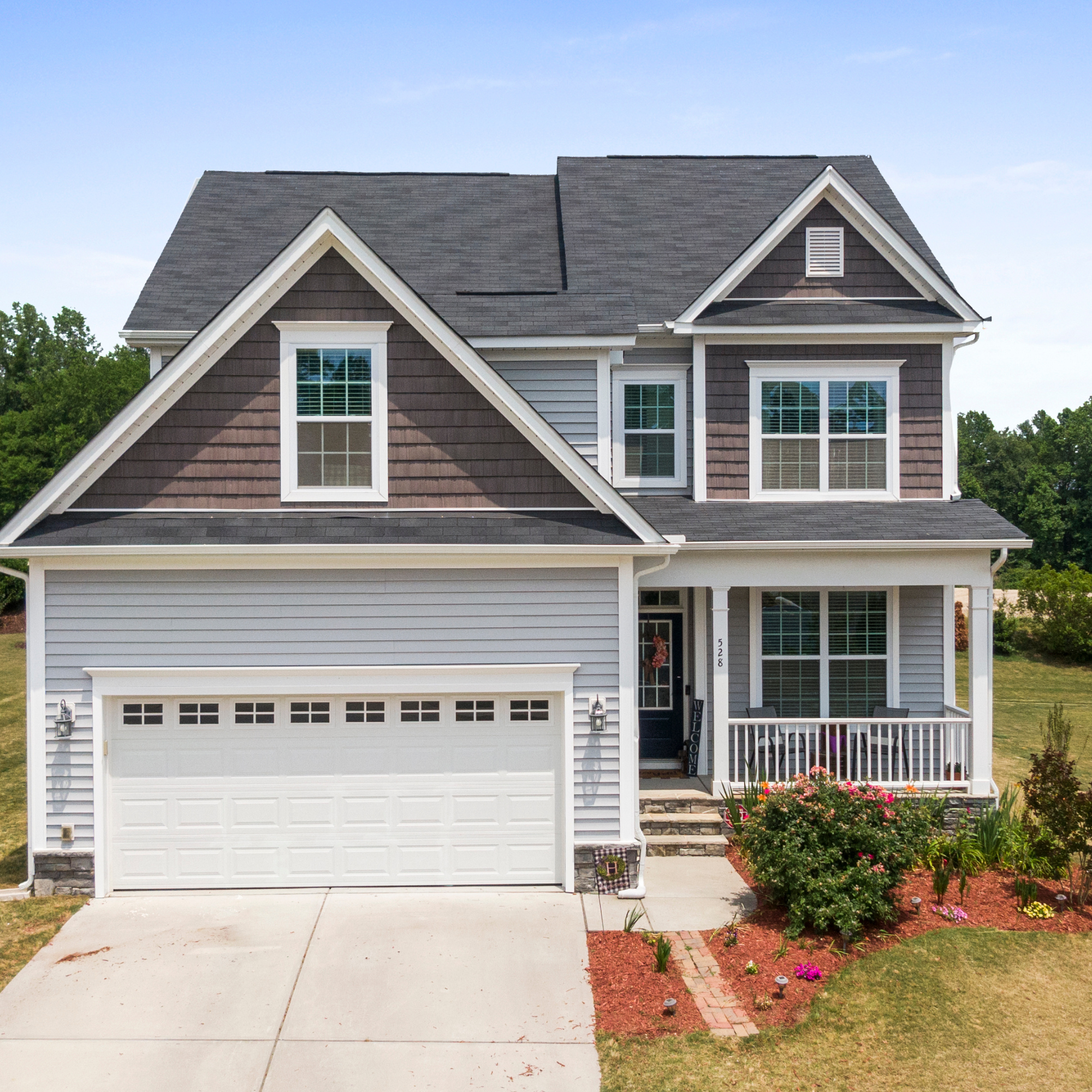 Light beige and brown house with multiple gabled roofs, stone and siding exterior, white window trims, a white garage door, and a small front porch with steps. The yard has some trees and shrubs, and the driveway is concrete.