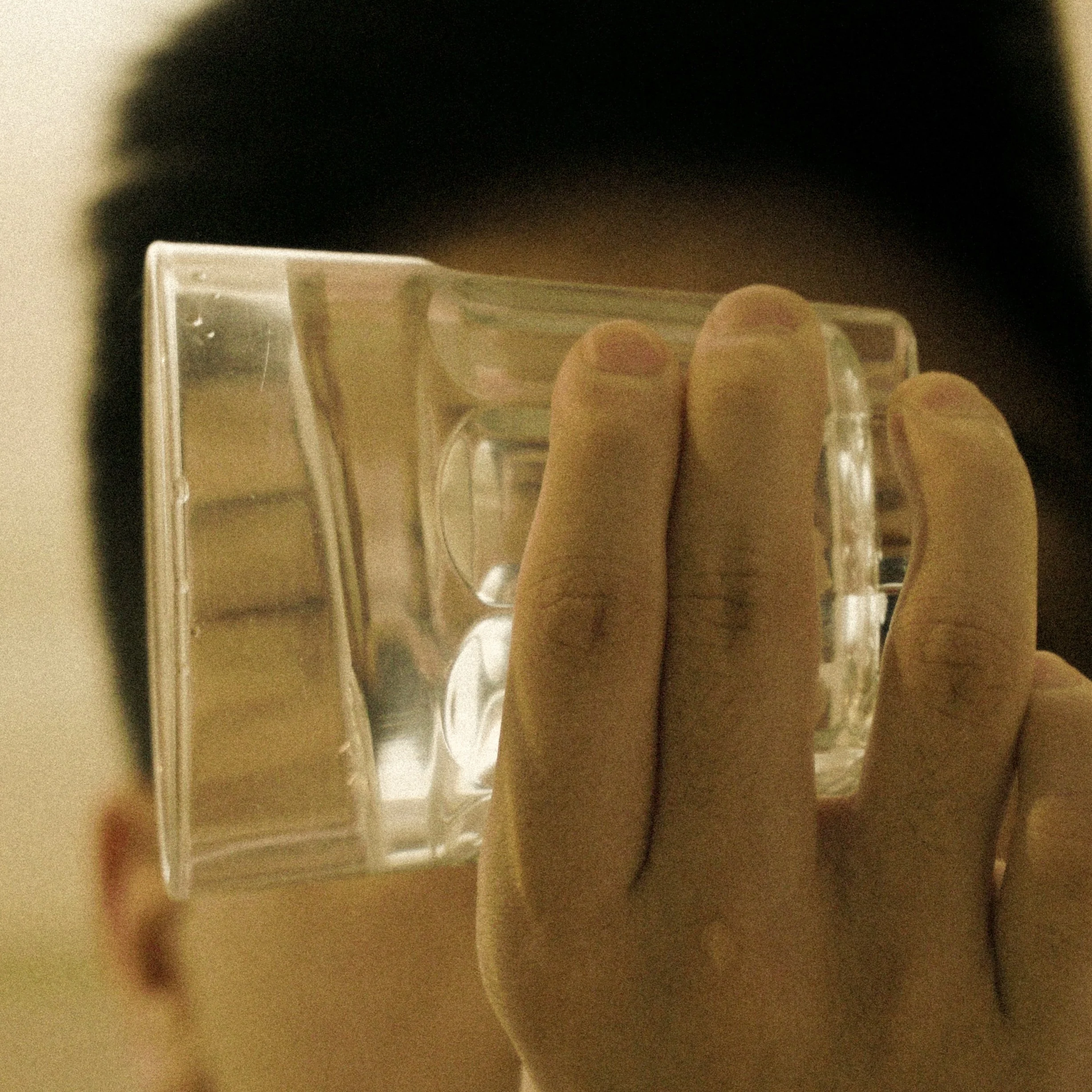 Two rectangular transparent objects resembling glasses or small containers held by a person's fingers, with a blurry background and a black area at the top of the image.
