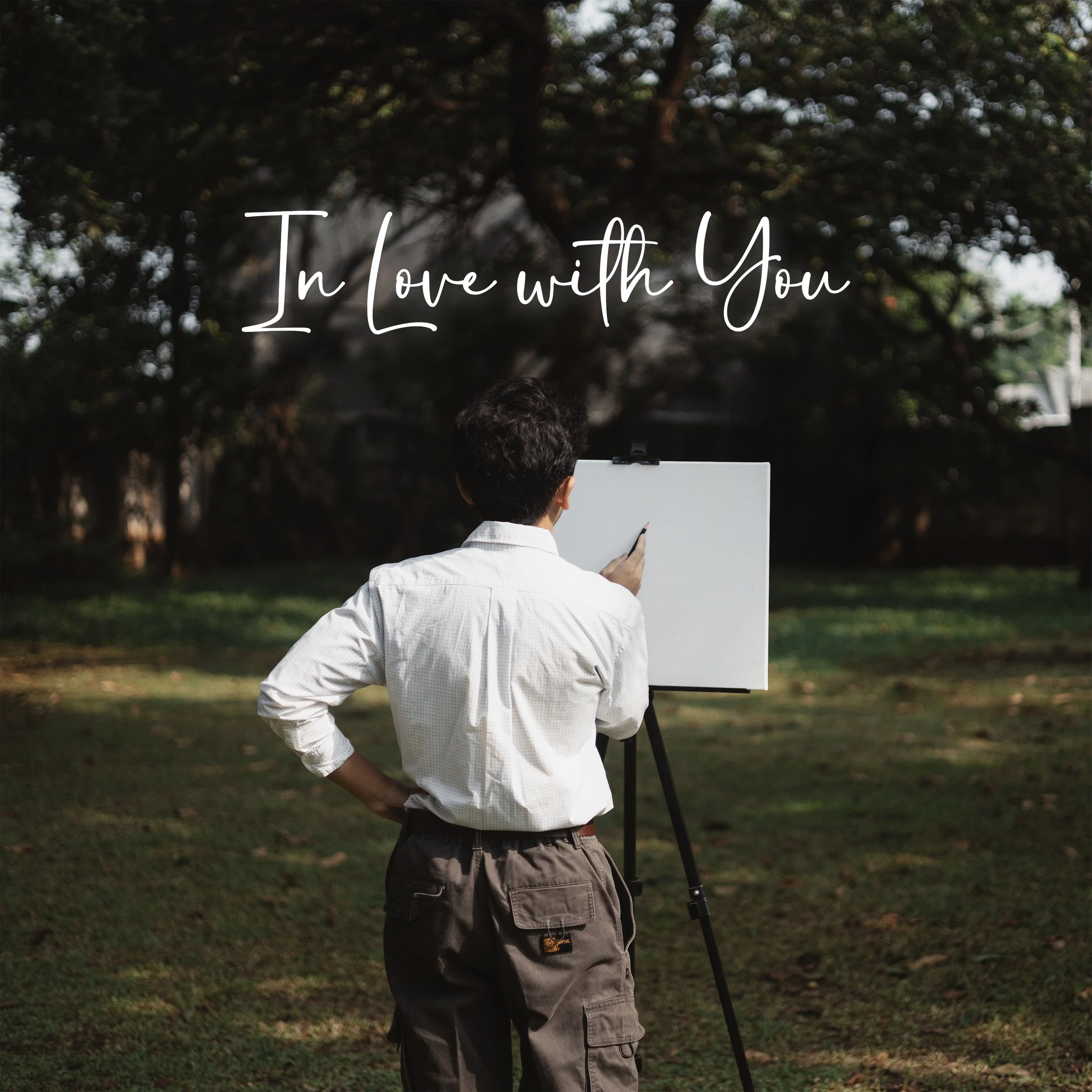A man in a white shirt and brown cargo shorts is standing outdoors, holding a white board and facing away from the camera. Behind him, there are trees and a cloudy sky. Text on the image reads "In Love with You" in white cursive font.