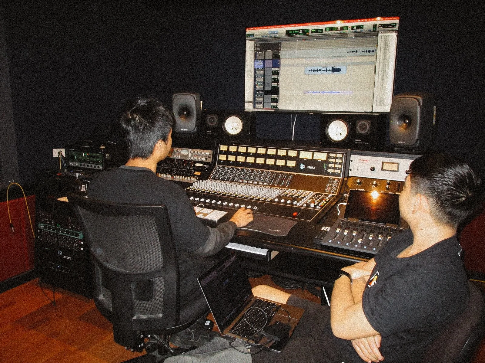 Two men working in a music recording studio, with a large mixing console, speakers, a computer screen displaying audio tracks, and various audio equipment.