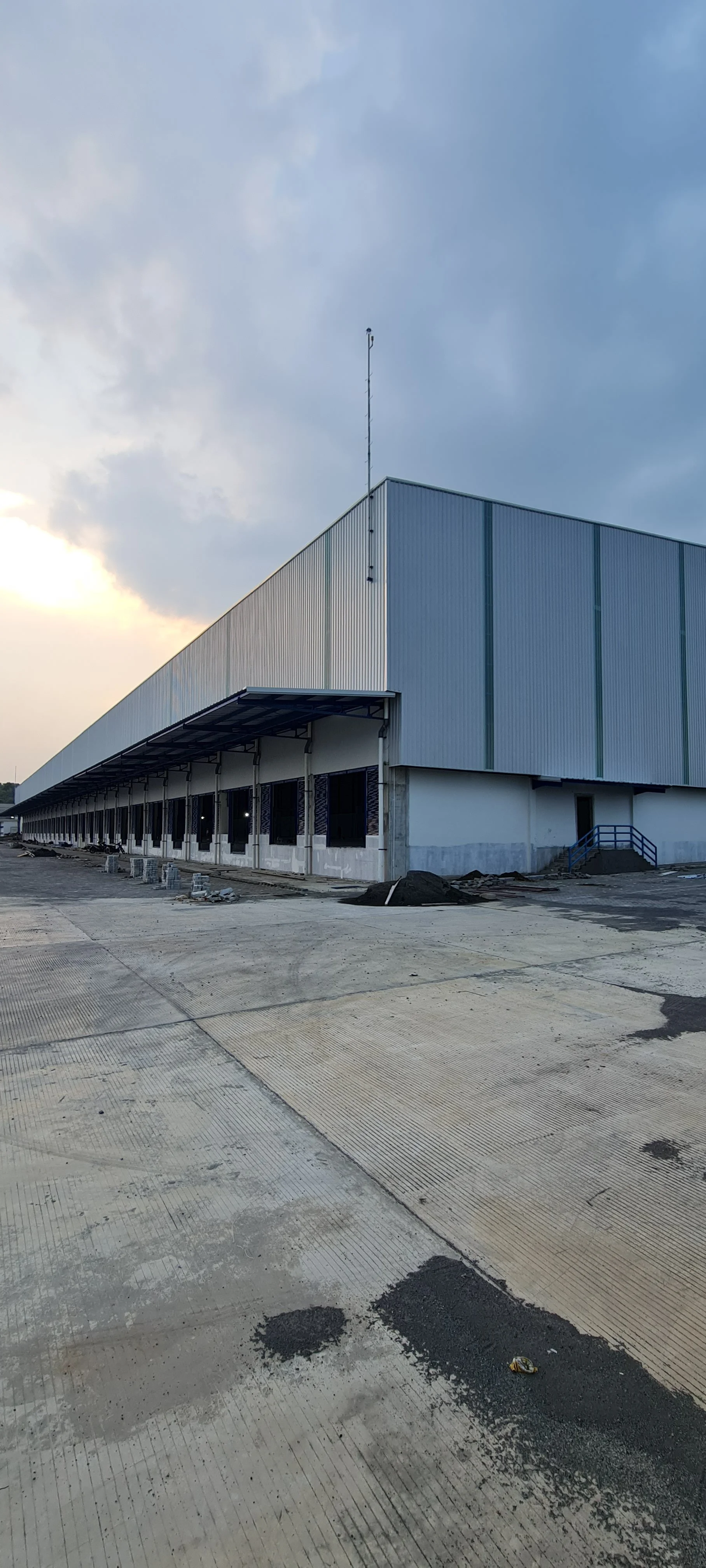 Exterior of a large industrial building under construction with a corrugated metal facade and multiple windows. The foreground shows a paved area with patches of asphalt, construction debris, and oil stains. The sky is partly cloudy with the sun setting in the background.