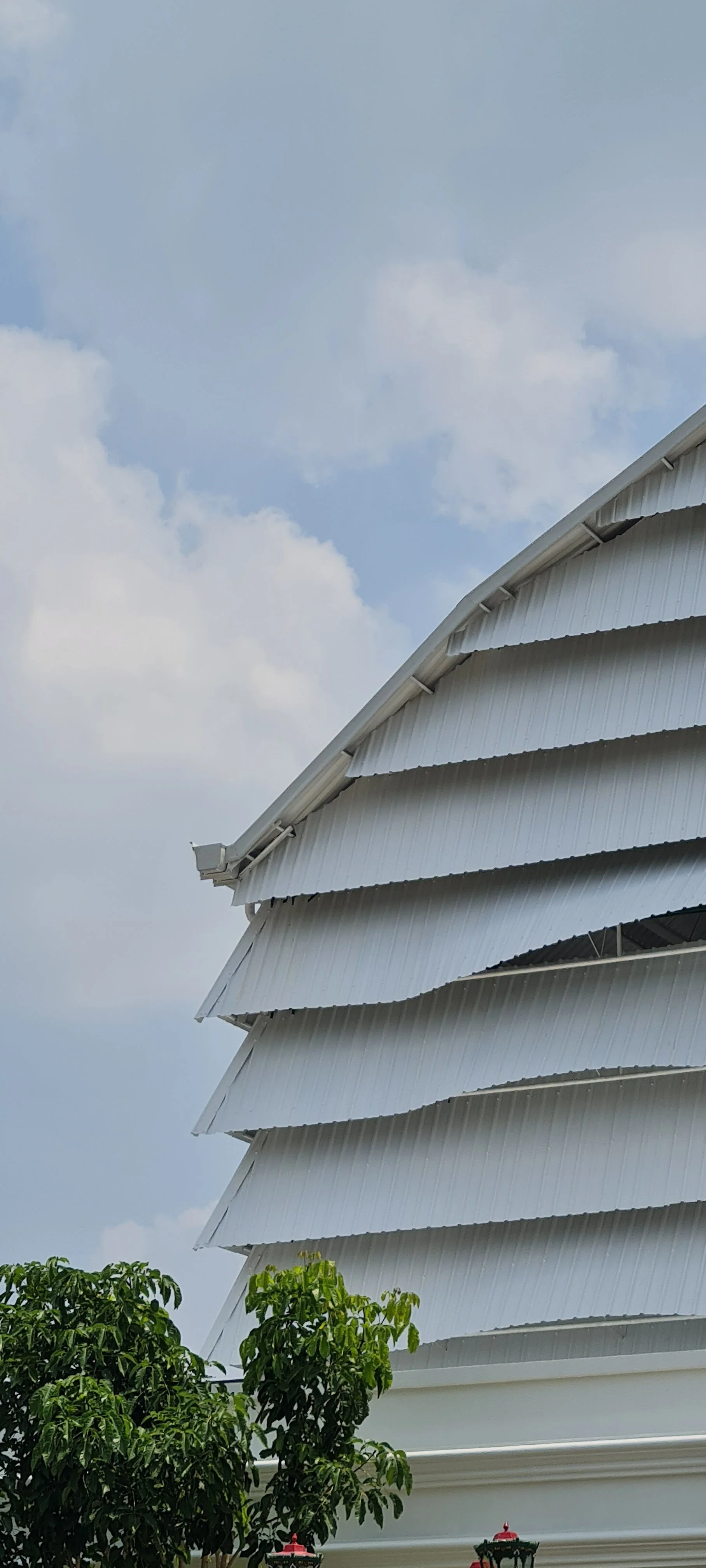 Close-up of a white, multi-layered, curved metal roof with a cloudy sky in the background and green trees in the foreground.
