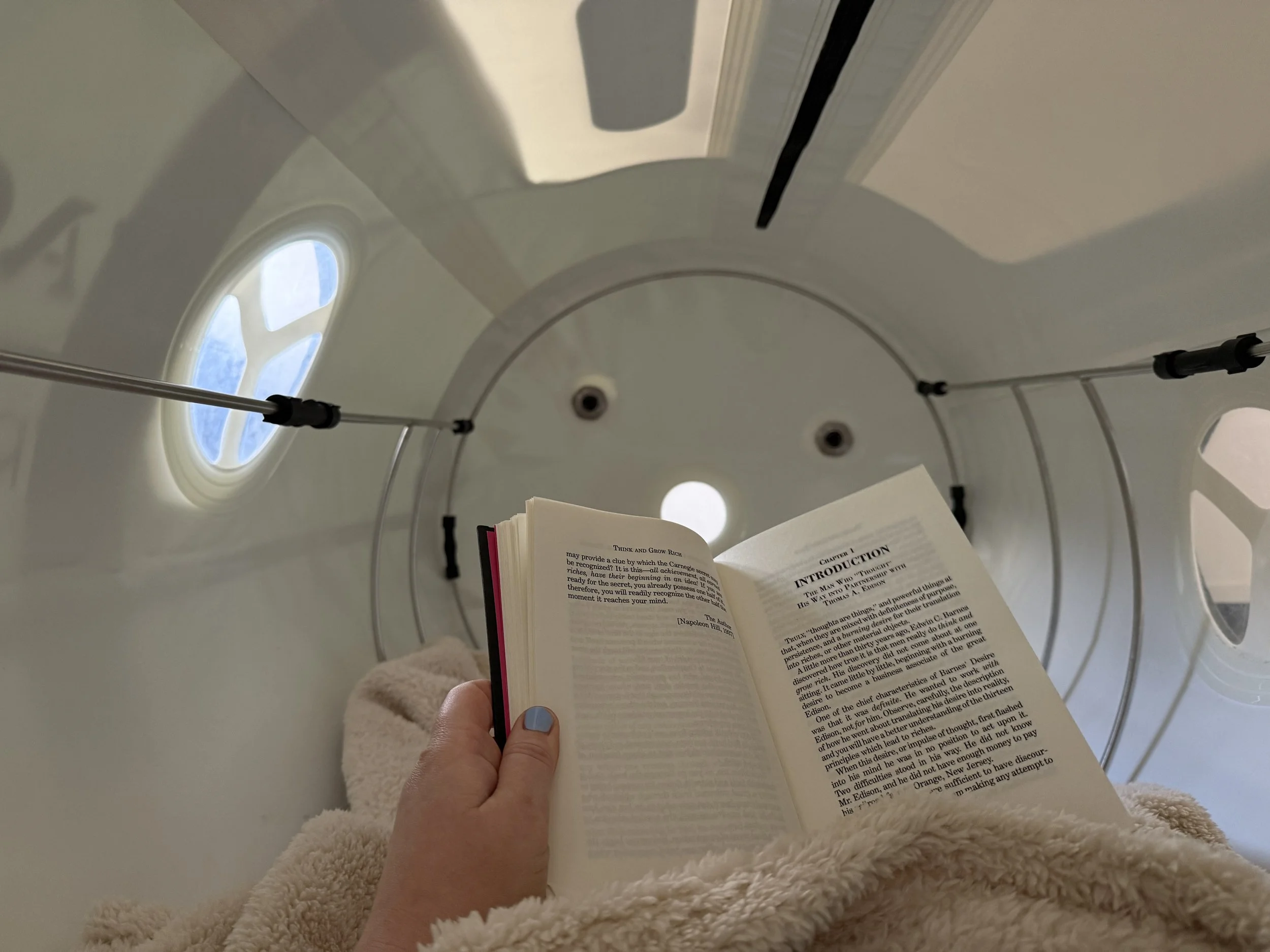 Person reading a book while lying on a plush blanket with their hand visible, inside a room with a round window and a ceiling with a curved white surface.