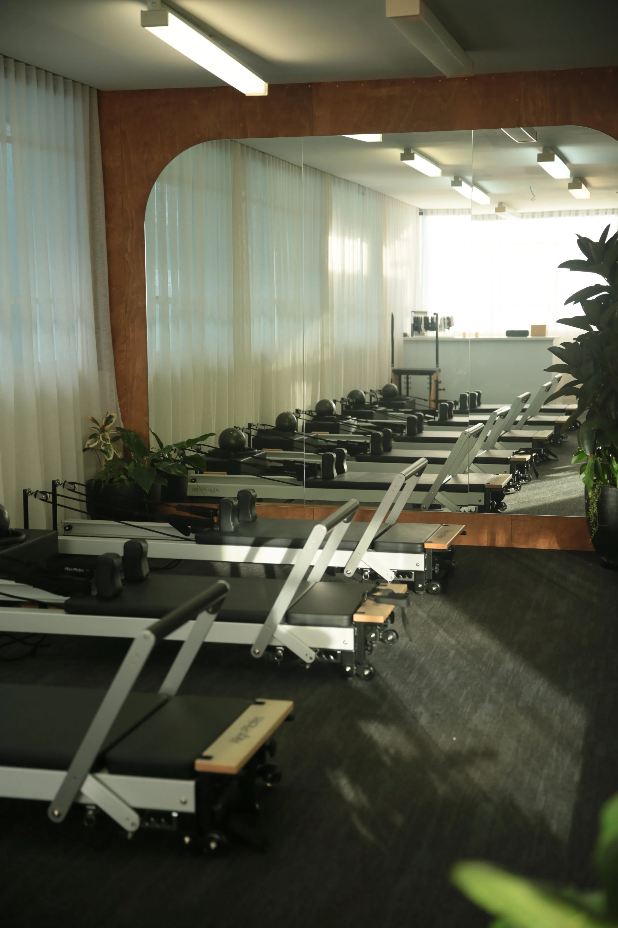 Row of Pilates reformer machines in a fitness studio with large windows and curtains, greenery in the foreground, and a mirrored wall.