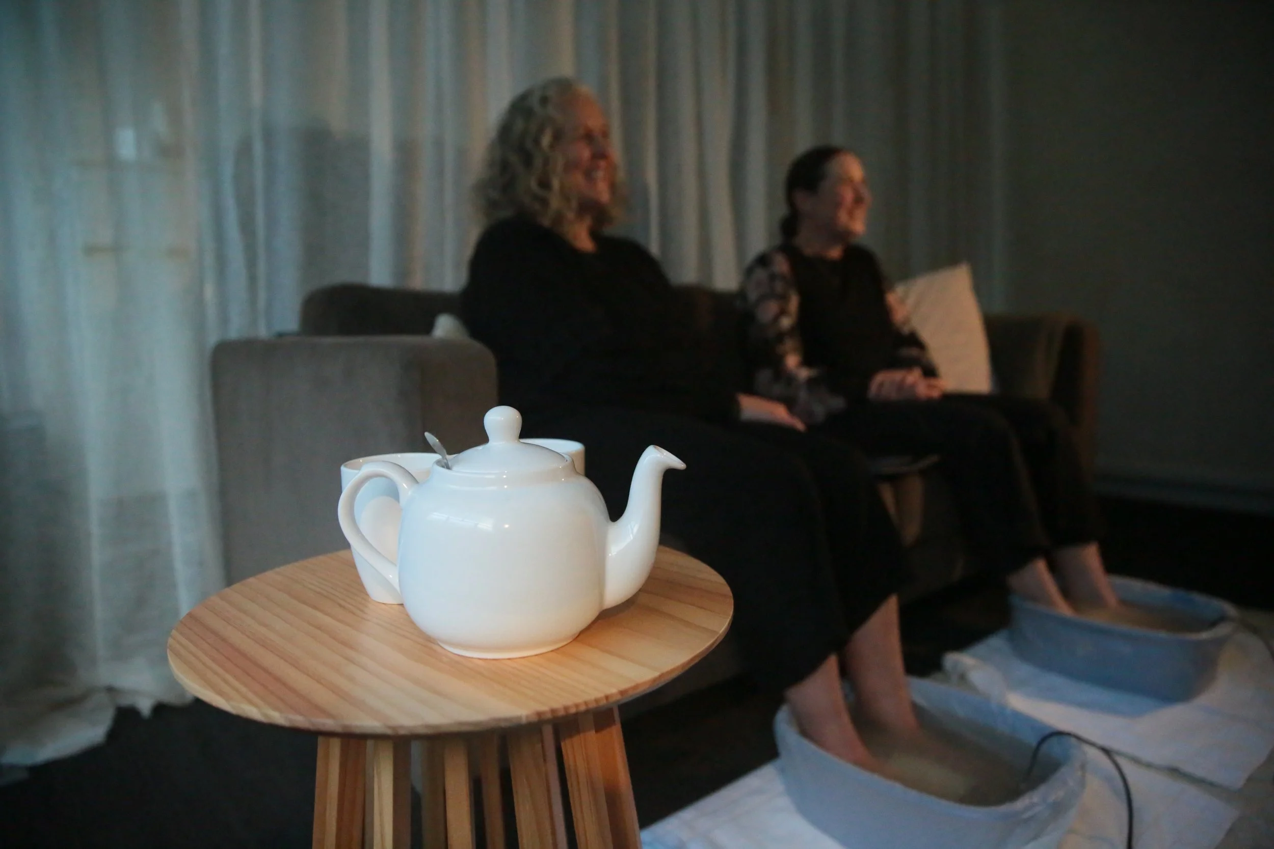 A white ceramic teapot with a lid and spout placed on a small round wooden table, with two women sitting on a gray couch in the background, one with curly gray hair and the other with dark hair tied back, in a softly lit room with curtains.