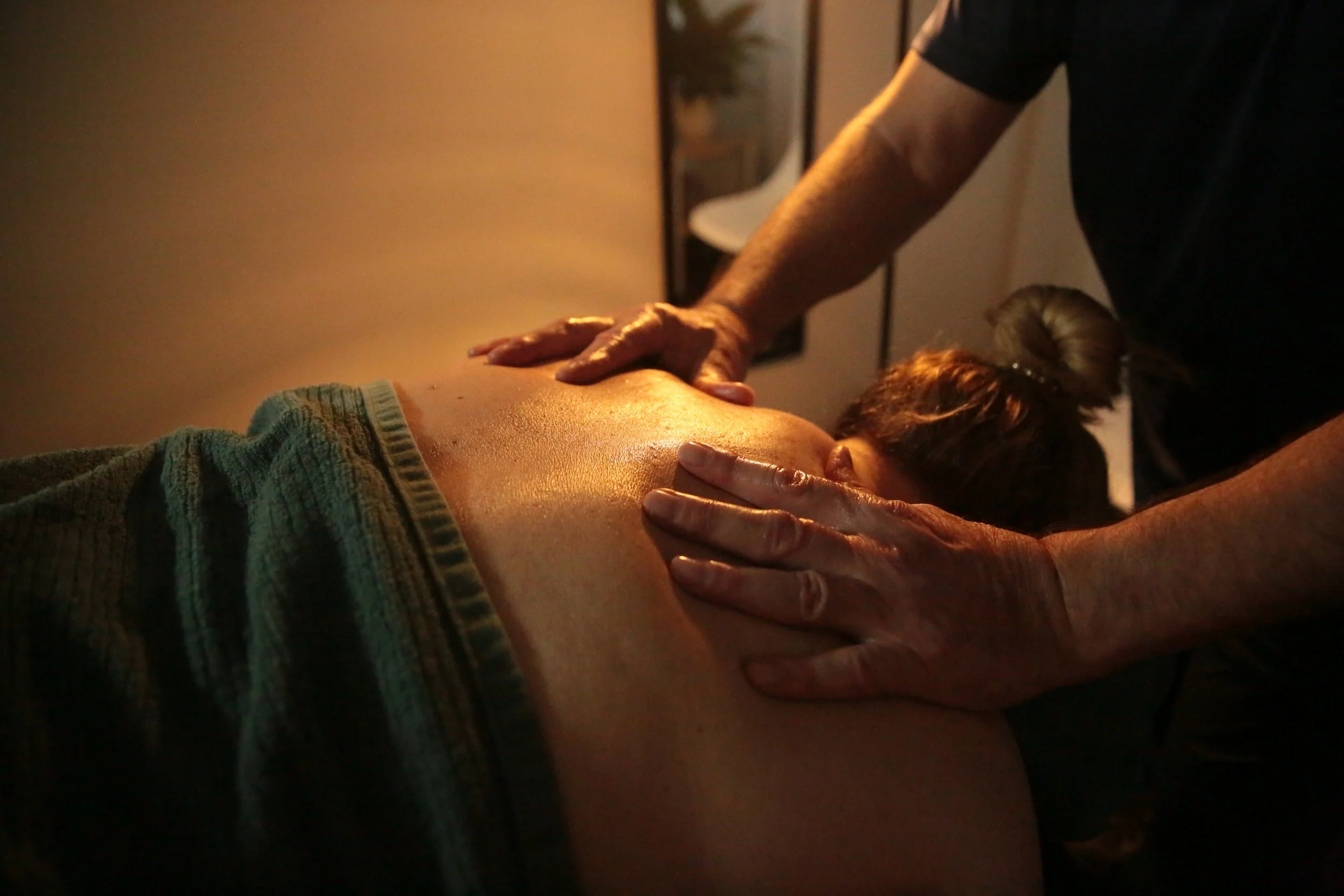 A person receiving a massage on their back, with the masseuse's hands applying pressure. The scene is dimly lit, suggesting a relaxing environment.