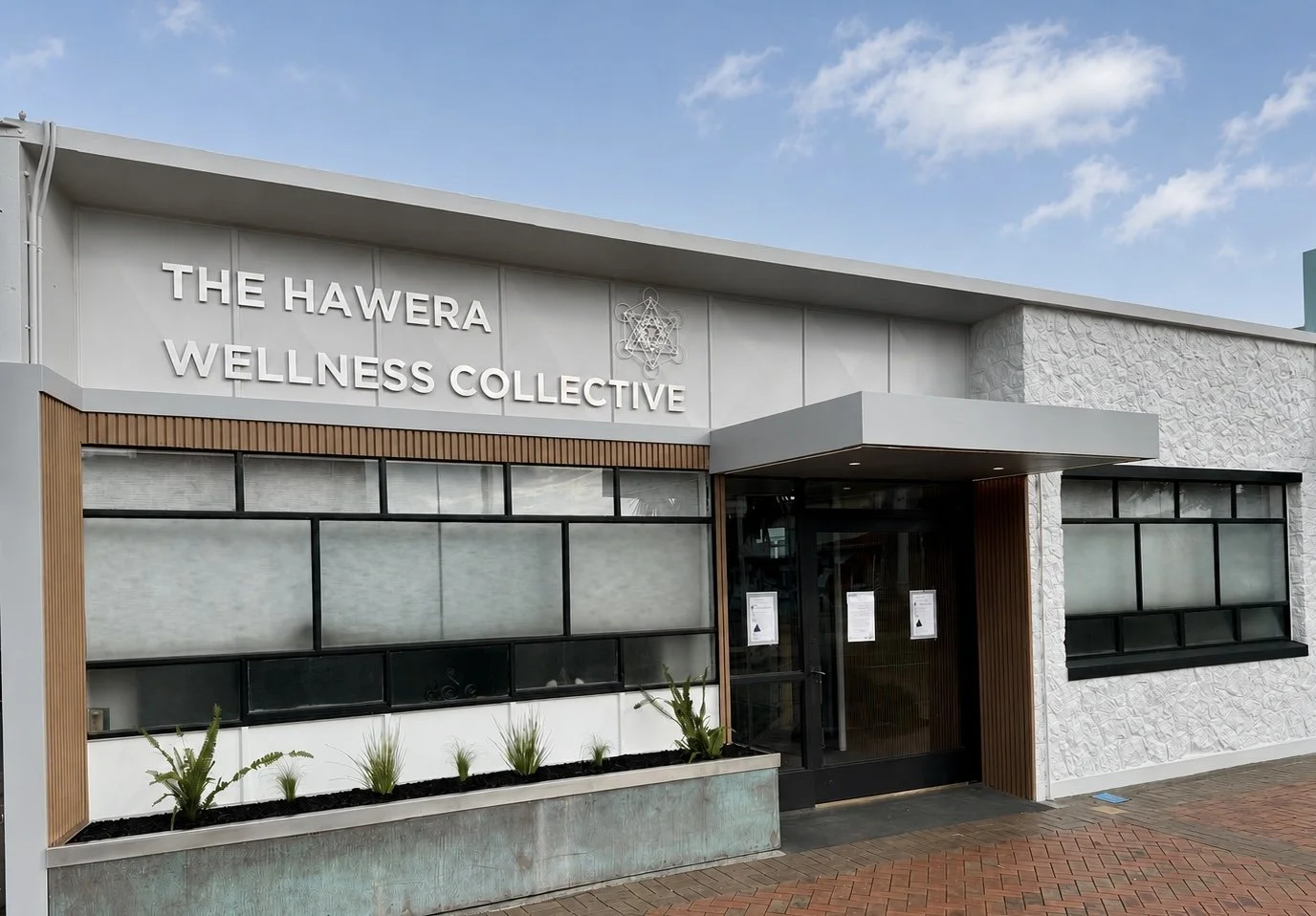 Front view of The Hawera Wellness Collective building with modern design, large windows, and a small garden planter in front, under a partly cloudy sky.