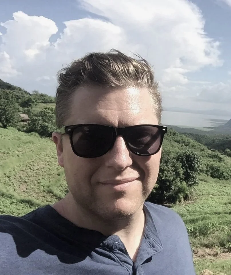 A man with light skin and short brown hair, wearing black sunglasses and a dark blue shirt, taking a selfie outdoors on a sunny day with a hilly green landscape and cloudy sky in the background.