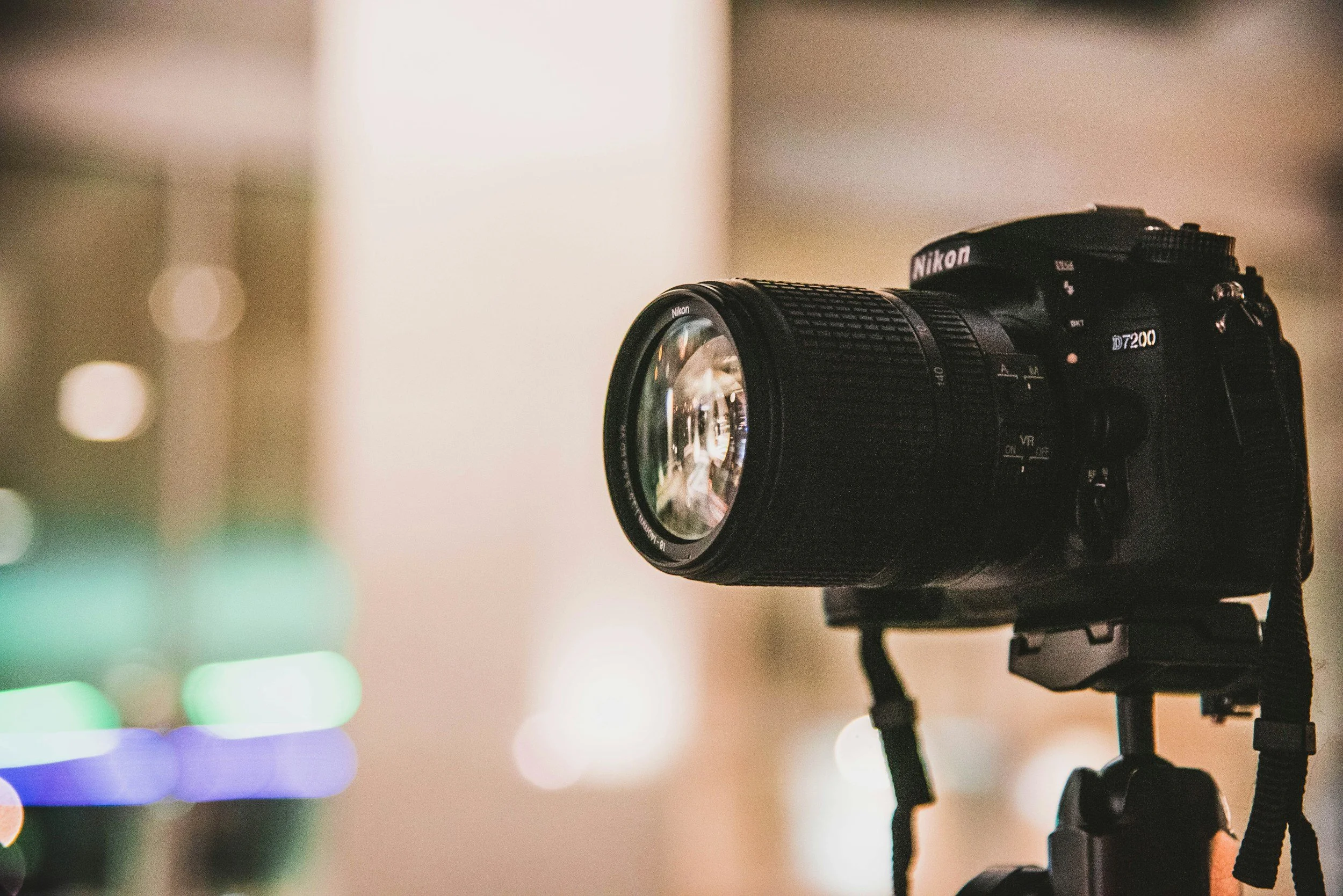 Close-up of a Nikon D7200 DSLR camera mounted on a tripod, with a blurred colorful background of lights.