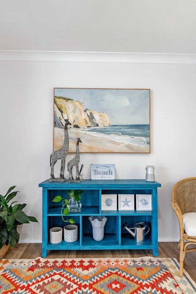 A white wall decorated with a large beach landscape painting, a blue console table with beach decor items, and a wicker chair on the right. There are also green plants and a red, beige, and gray patterned rug on the floor.