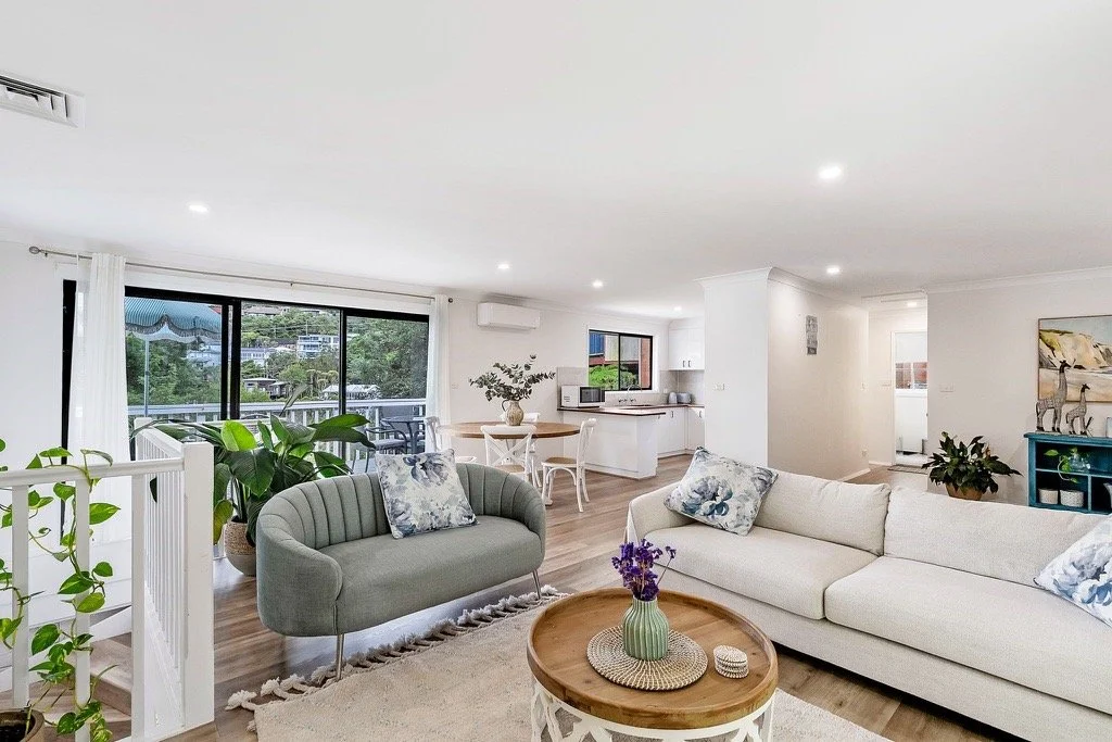 Open-concept living room with white walls, large window, gray and white sofas, wooden coffee table with decorative items, potted plants, and view of the balcony with outdoor furniture.