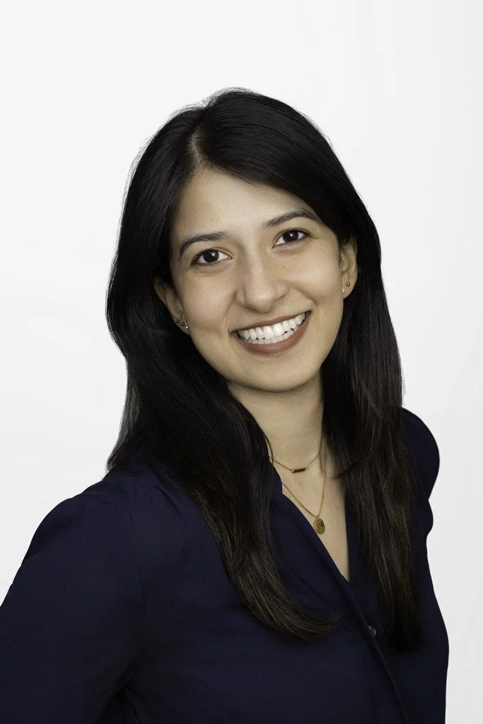 Young woman with long dark hair smiling at the camera, wearing a dark blue blouse and gold jewelry against a plain white background.