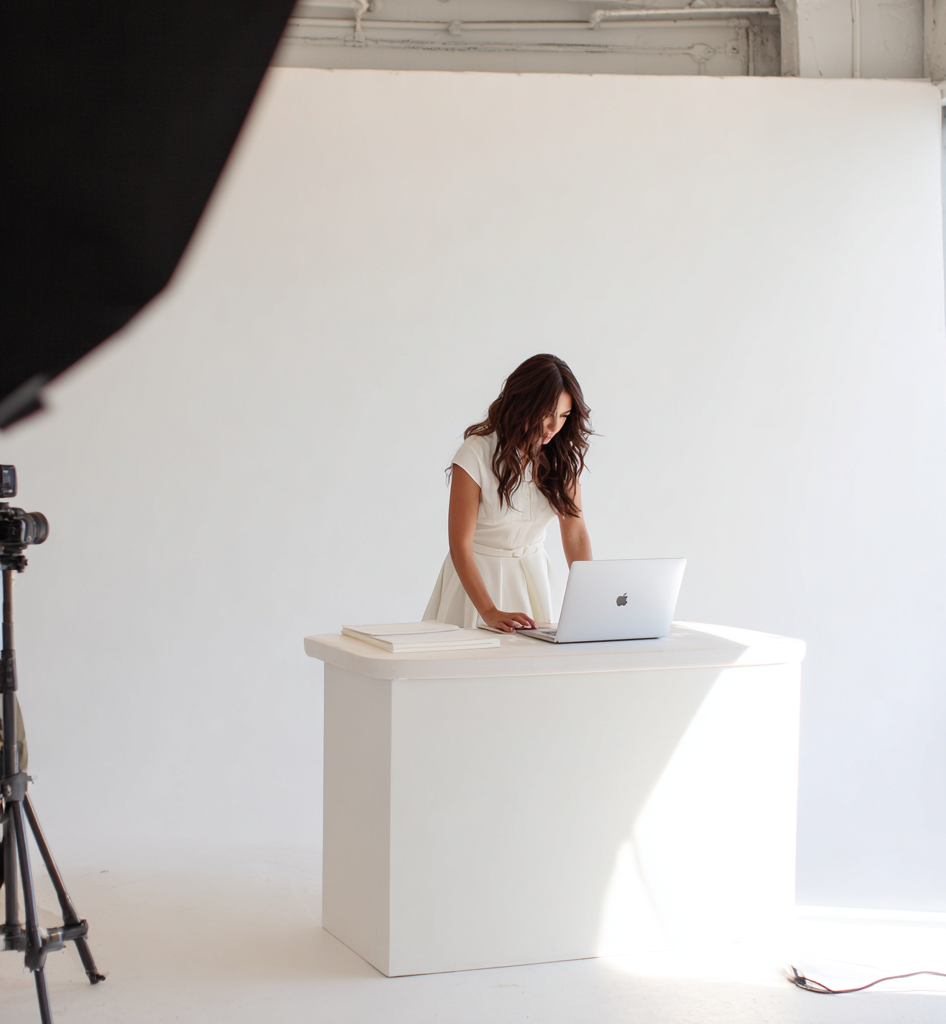 A woman standing at a white table with a laptop and notebooks in a photo studio with white walls, with camera equipment visible.