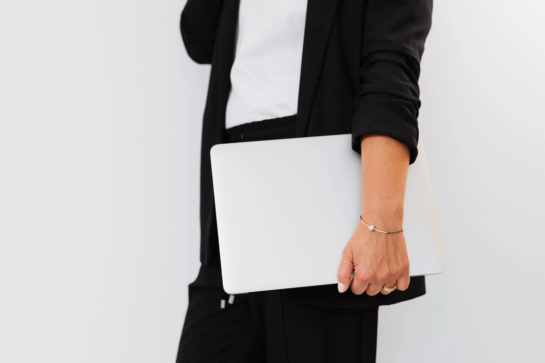 Person dressed in black blazer and pants holding a closed silver laptop, wearing a bracelet and ring on their hand, against a plain light-colored background.