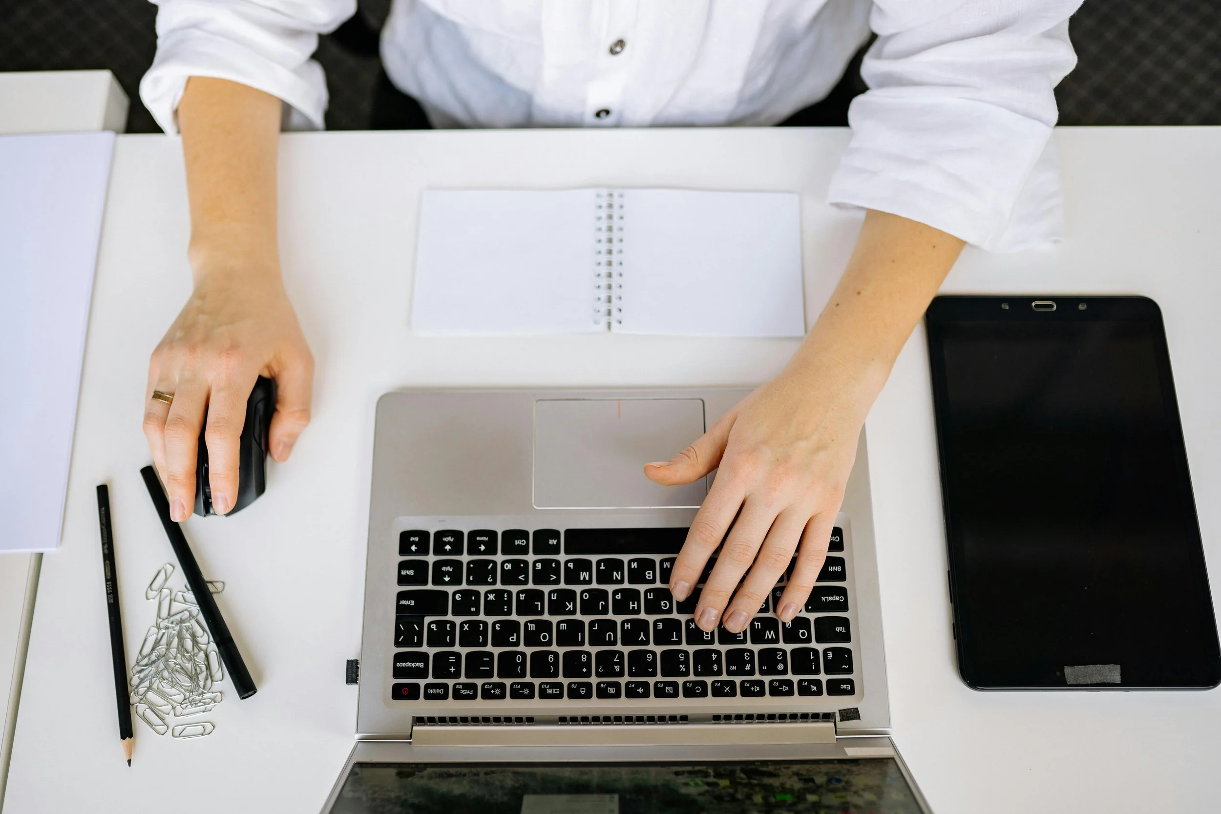 A person using a laptop computer at a white desk, with a smartphone, a notebook, papers, paper clips, pencils, and pens around.