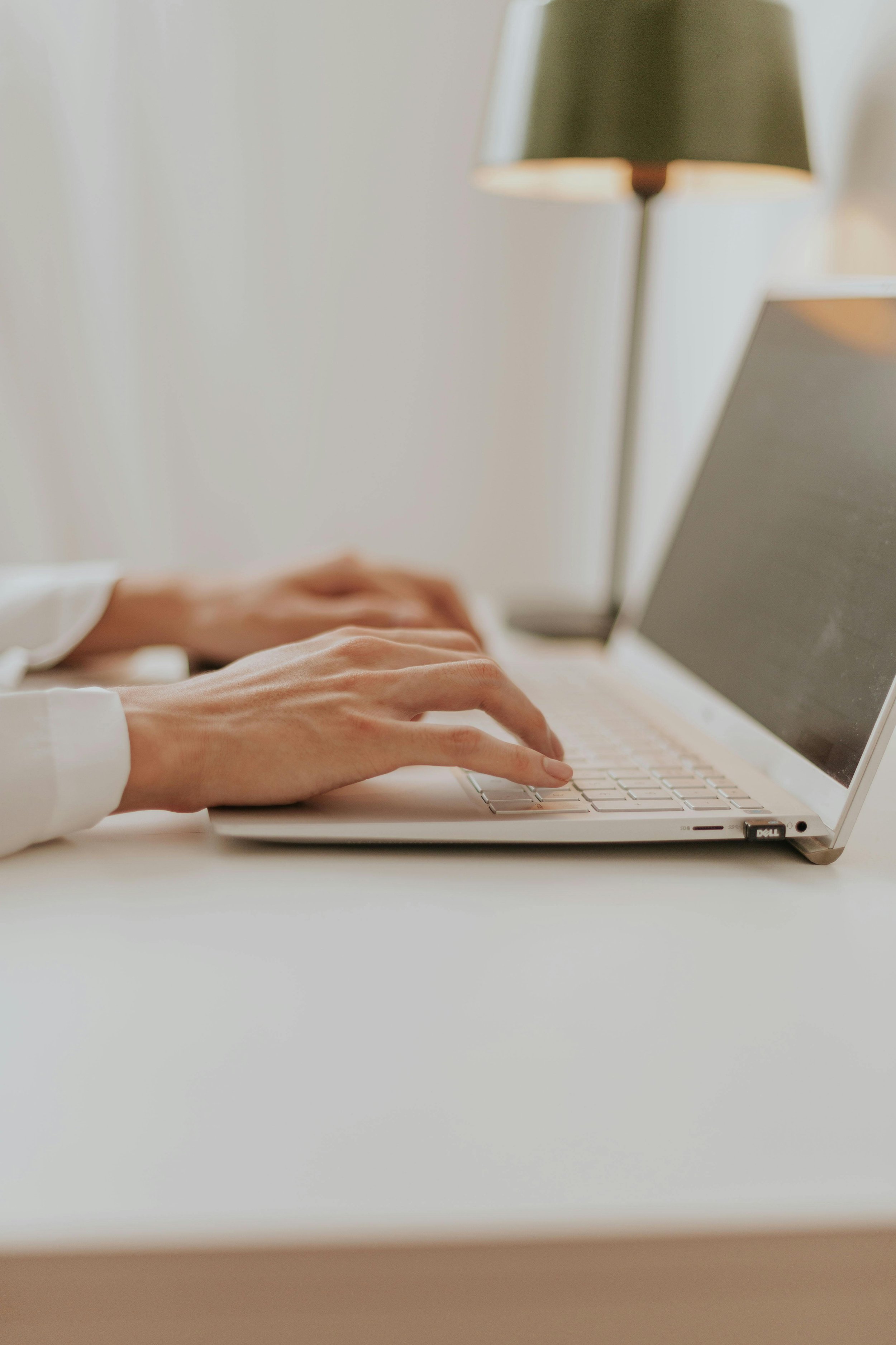 Person typing on a white laptop keyboard, with a white desk and a green lampshade in the background.