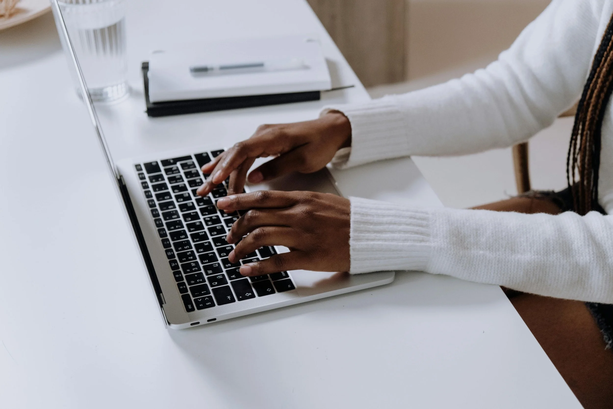 A person with dark skin and long braided hair typing on a silver laptop keyboard, wearing a white sweater, with a closed notebook and pen placed on a white table.