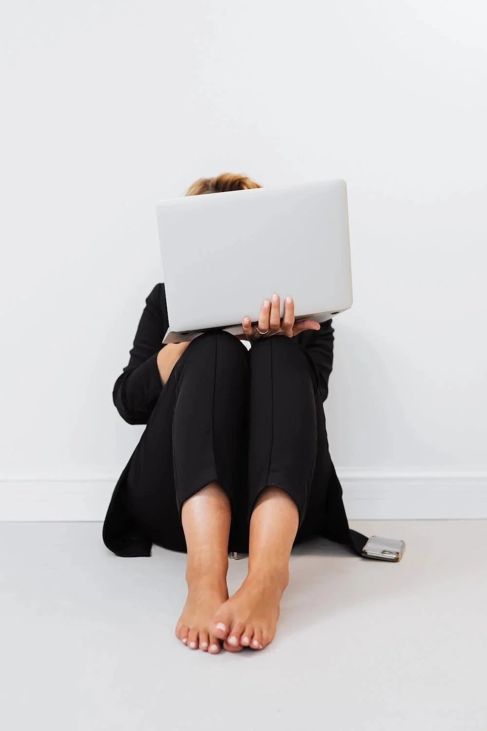Person sitting on the floor with knees drawn up, holding a closed laptop in front of their face, against a plain white wall.