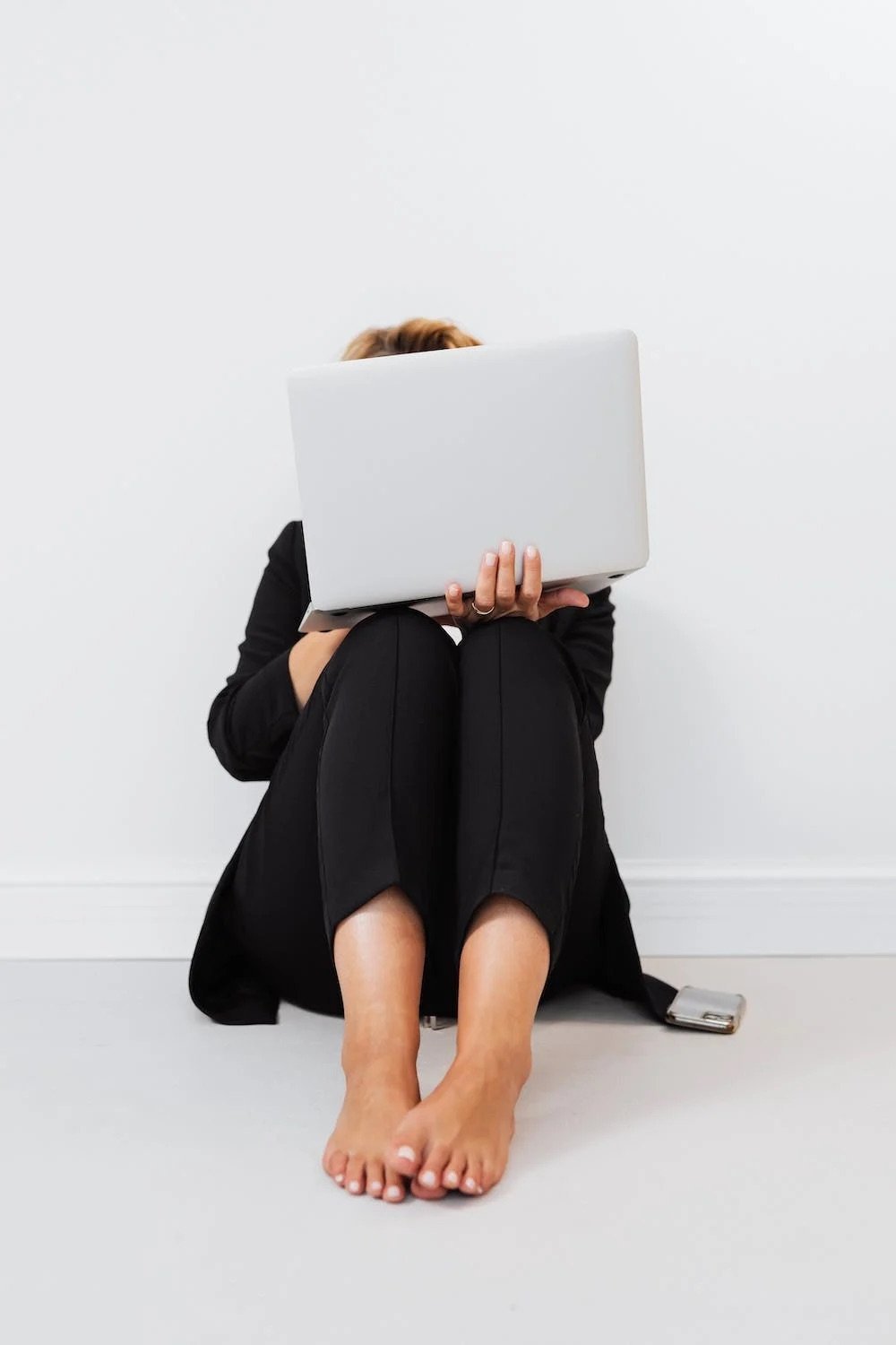 Person sitting on the floor with legs crossed, holding a laptop in front of their face, wearing a black outfit, in a minimalistic room with white walls.