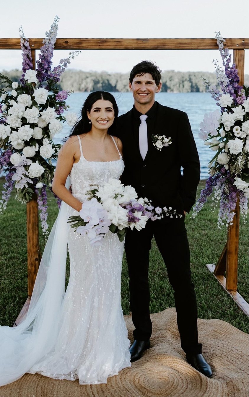 A bride and groom standing under a wooden wedding arch decorated with white and purple flowers, near a body of water with land in the background. The bride is wearing a white lace wedding dress and holding a bouquet, and the groom is dressed in a black suit with a white tie and boutonniere.